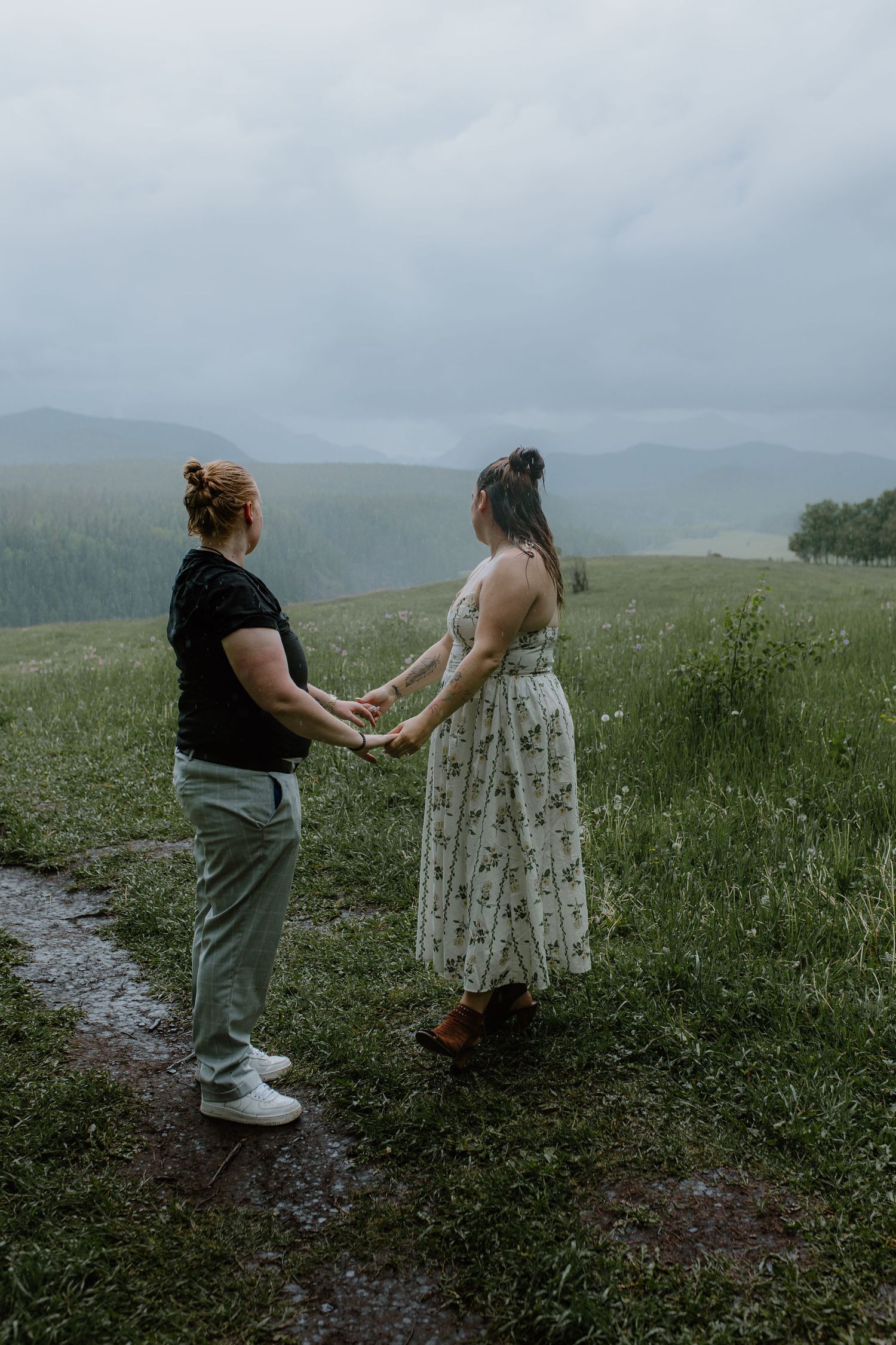 A couple holding hands on a muddy path surrounded by green fields and misty mountains.