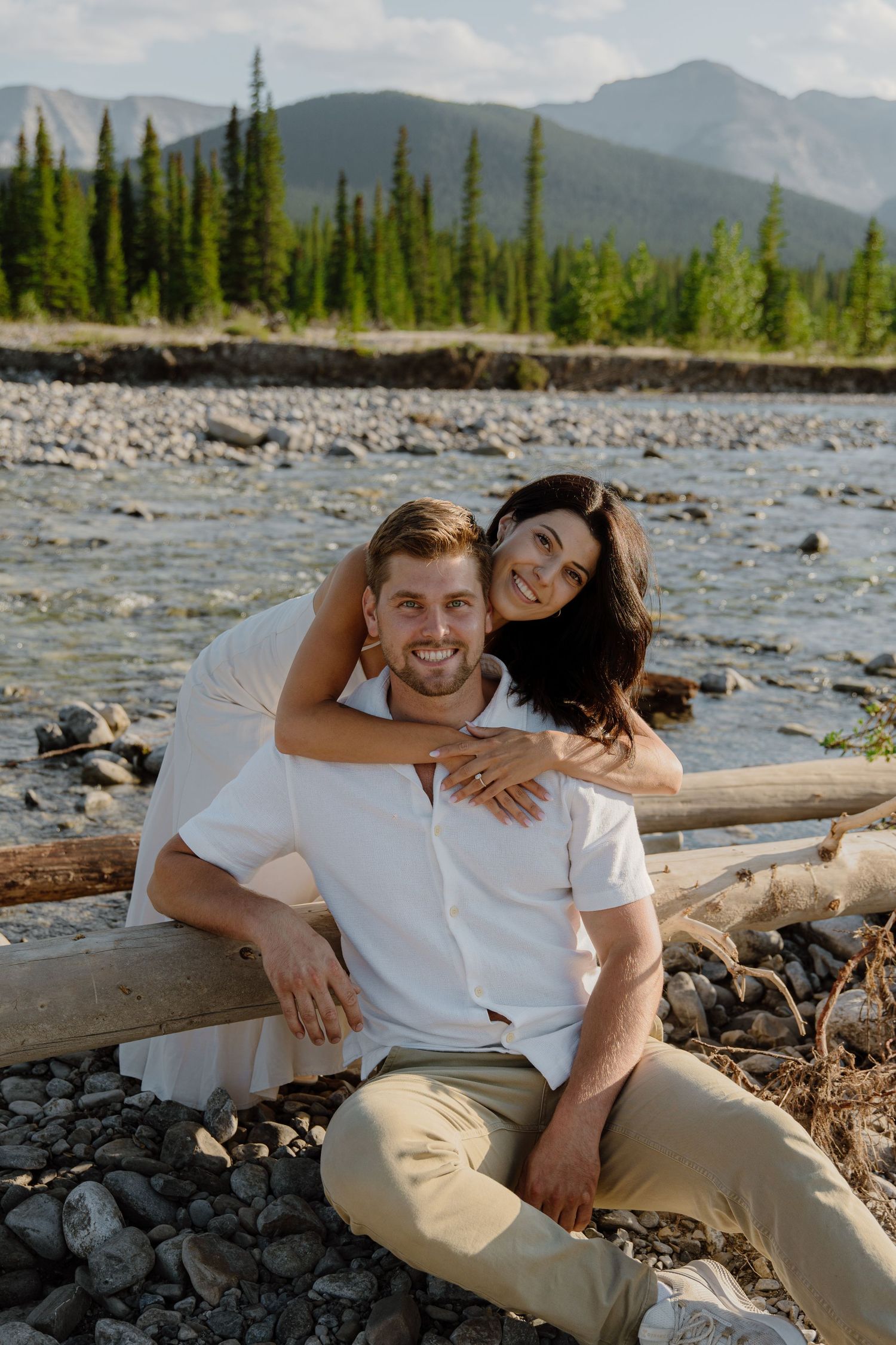 Couple posing by a rocky river with pine trees and mountains in the background on a sunny day.