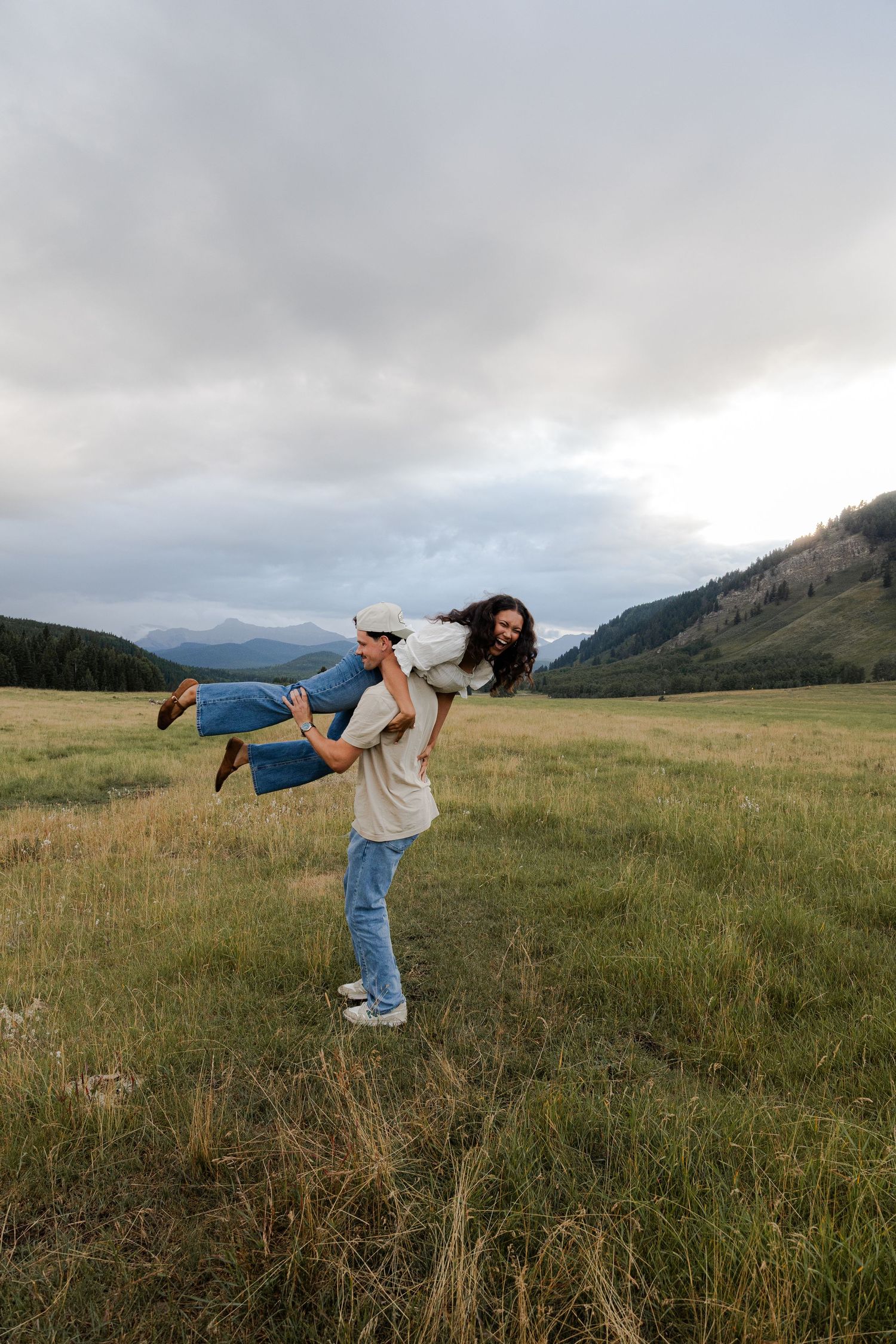 A man carries a woman on his back in a scenic mountain meadow under a cloudy sky.