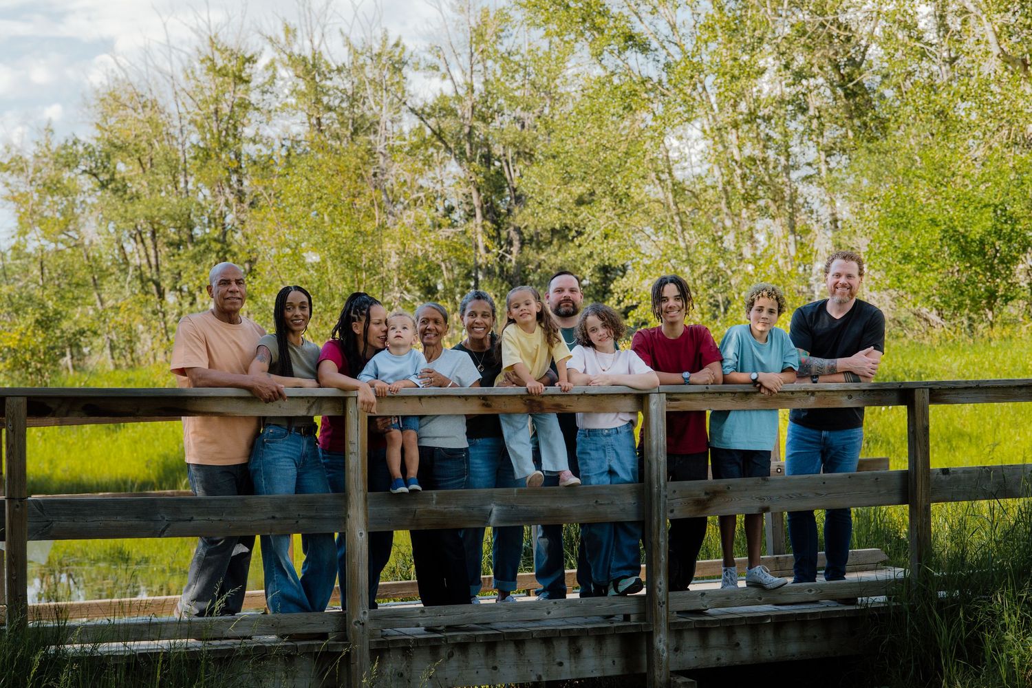 Large family group smiling together on a wooden bridge surrounded by lush green trees and wetlands.