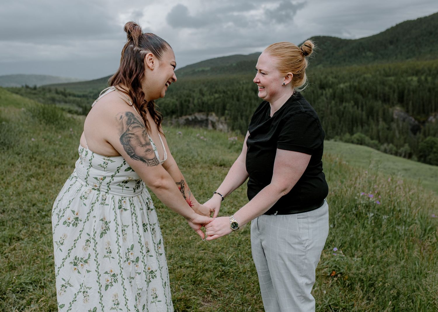 Two women hold hands and laugh together in a lush green mountain meadow during an outdoor couples photo session.