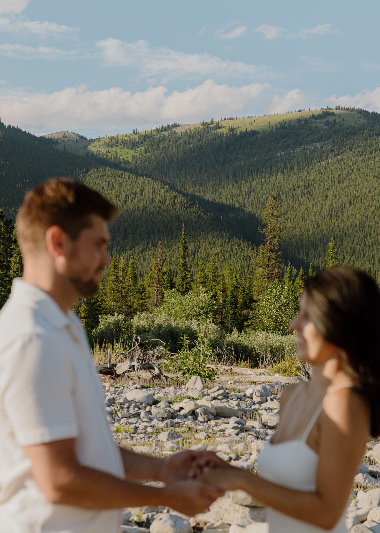 Couple holds hands during mountain elopement ceremony with lush green forest and rocky landscape backdrop in summer.