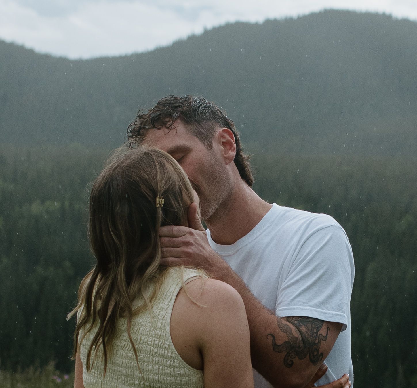 Couple sharing a romantic kiss outdoors with misty mountain forest backdrop on a rainy day.