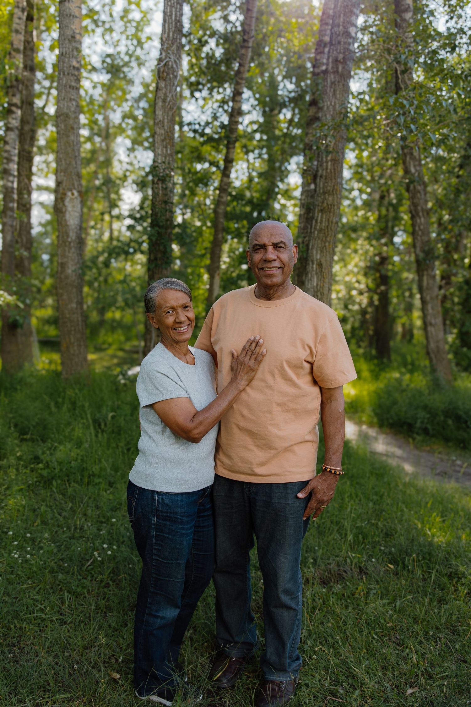 An elderly couple smiles warmly while standing together on a forest path surrounded by lush green trees.