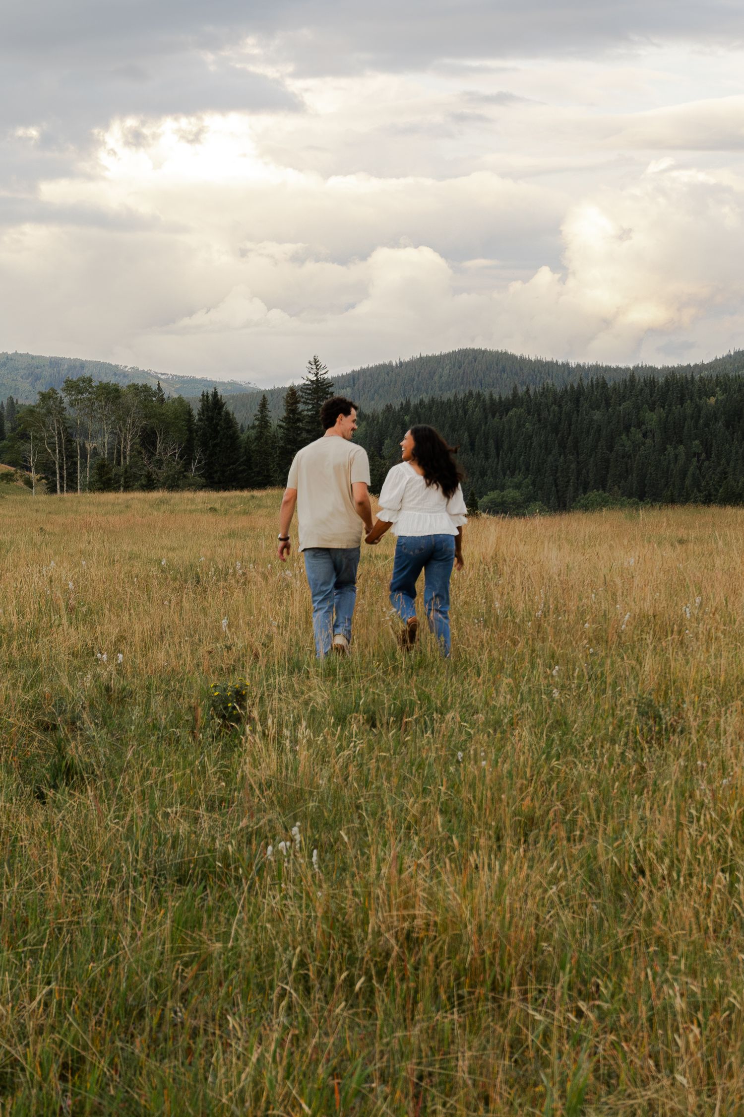 Couple holding hands walking through golden meadow with pine forest and cloudy mountain backdrop.