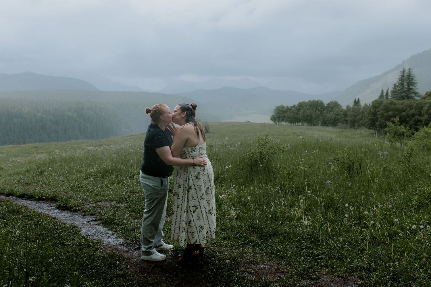 Couple sharing a romantic kiss in a lush green meadow with misty mountain views in the background.