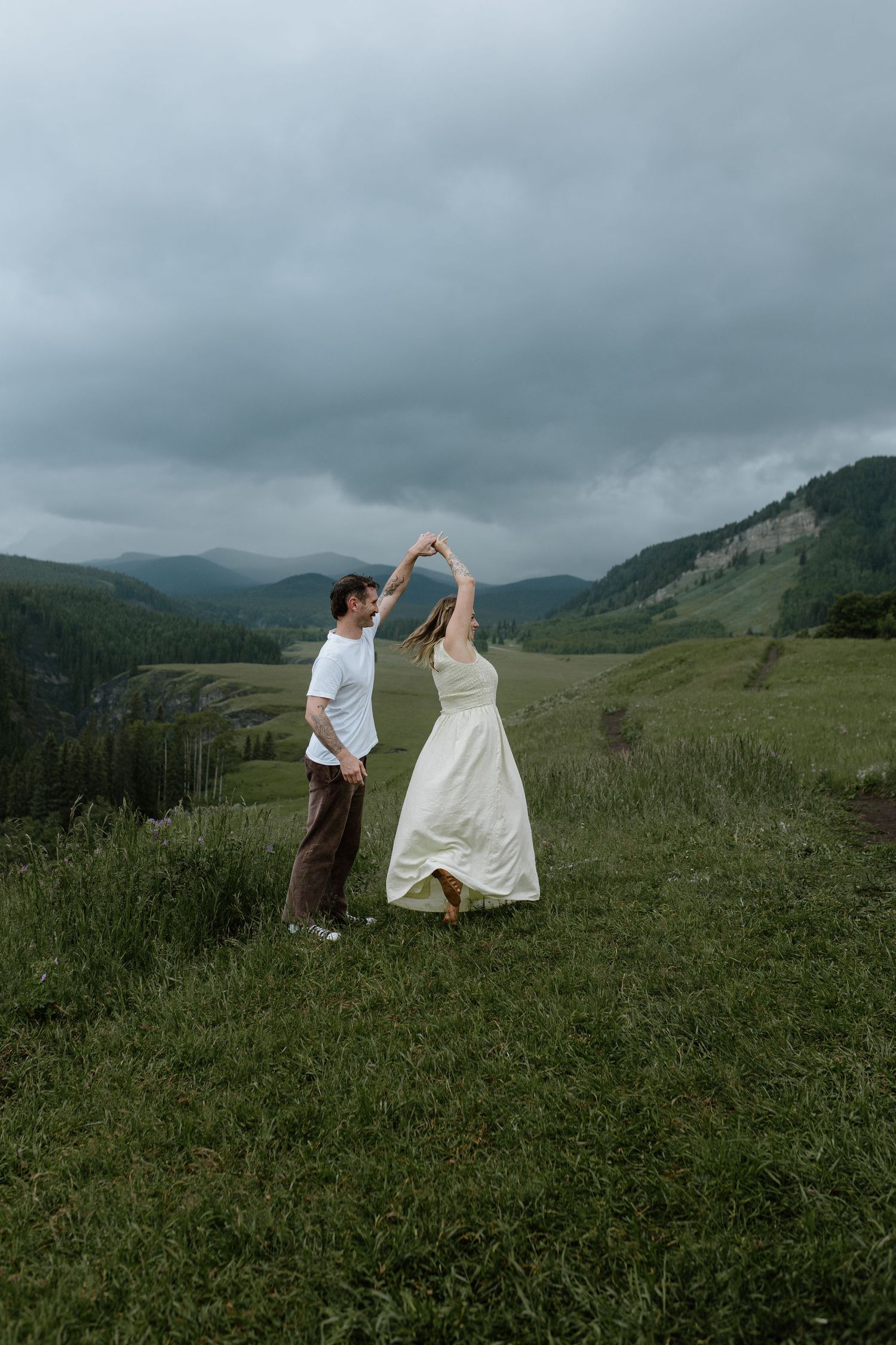 Couple dancing in a green mountain meadow, bride in white dress twirling under groom's hand with cloudy skies.