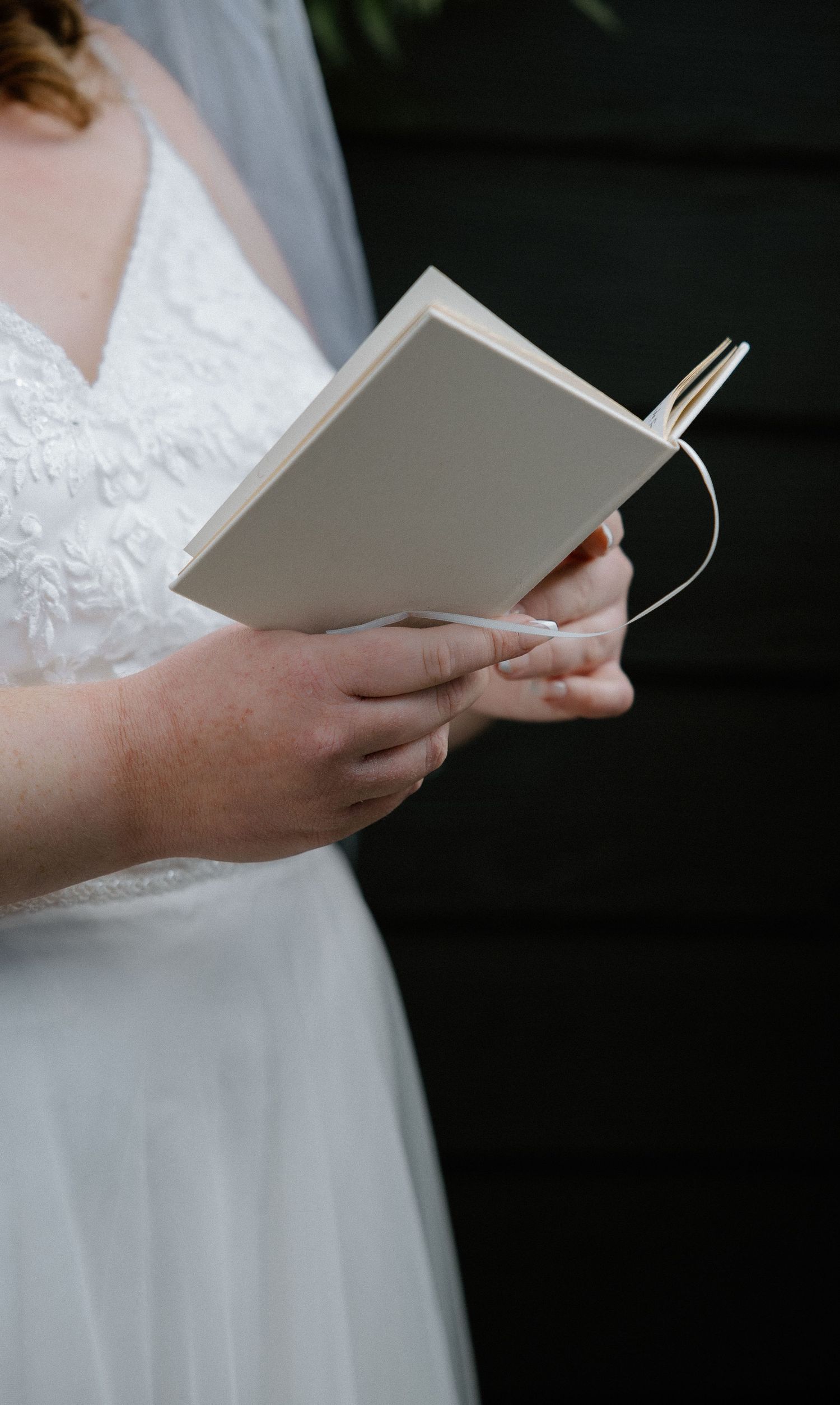 Bride in lace wedding dress and veil reads vows from small white booklet outdoors.