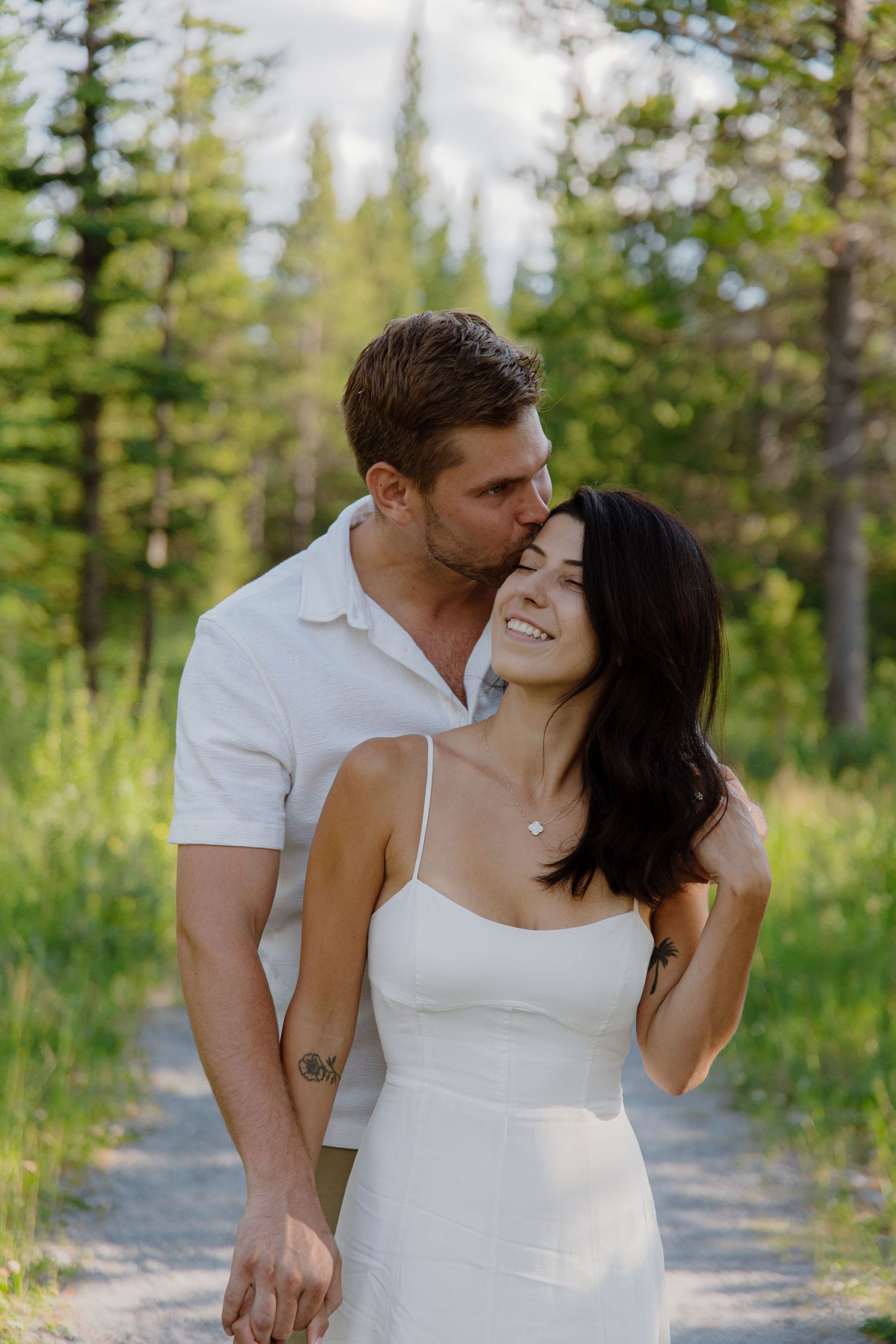 A couple shares a romantic moment on a forest path, man in white shirt kissing woman's temple as she smiles joyfully.