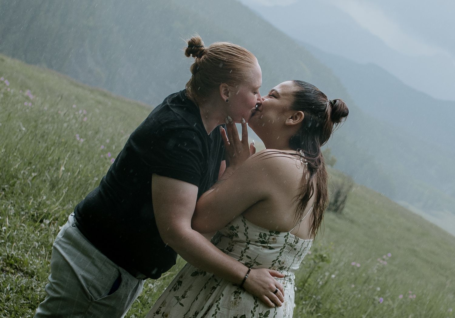 Two women sharing a romantic kiss in a scenic mountain meadow with misty peaks in the background.