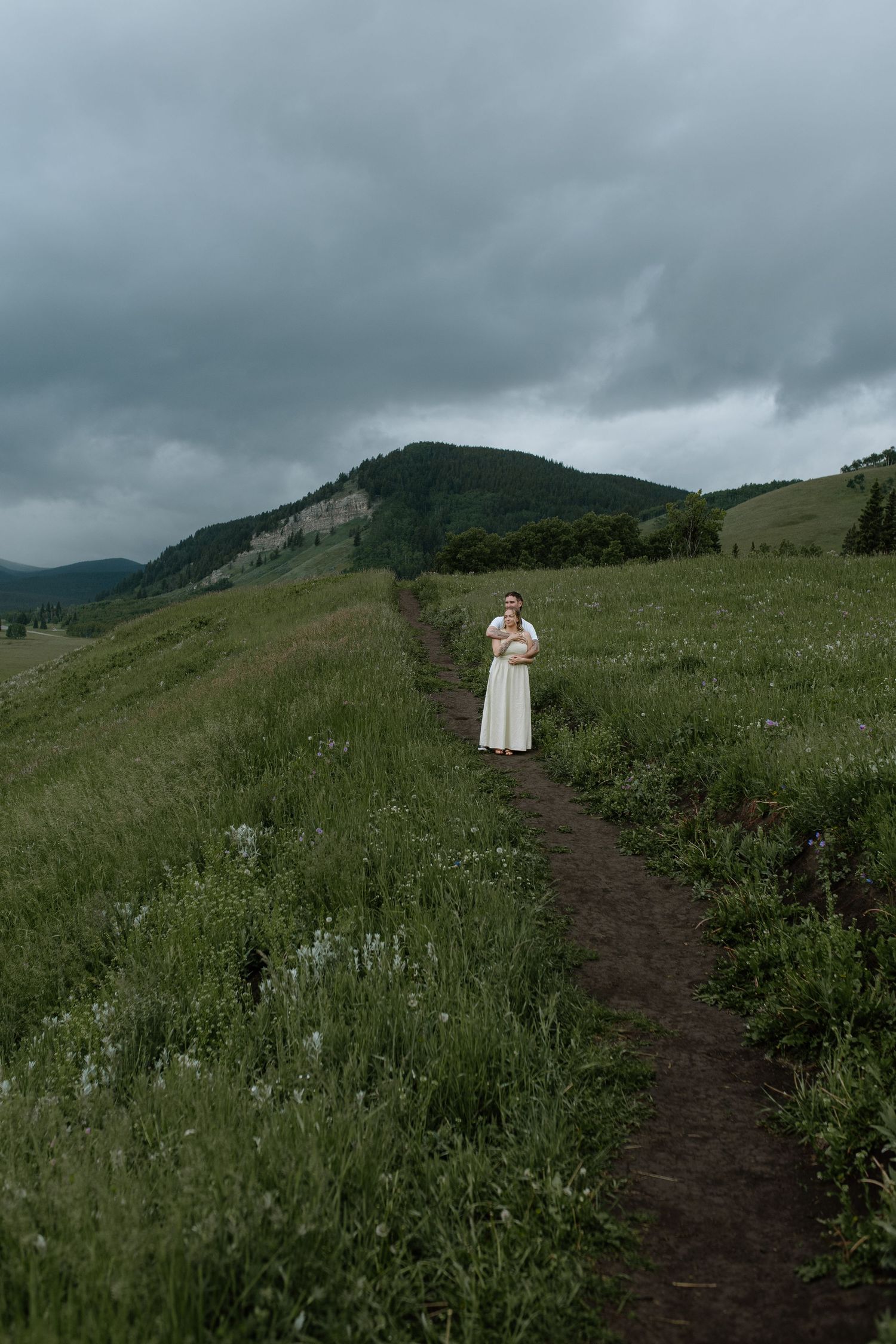 Woman in white dress holding flowers on a dirt path through green meadows with misty mountains under cloudy skies.