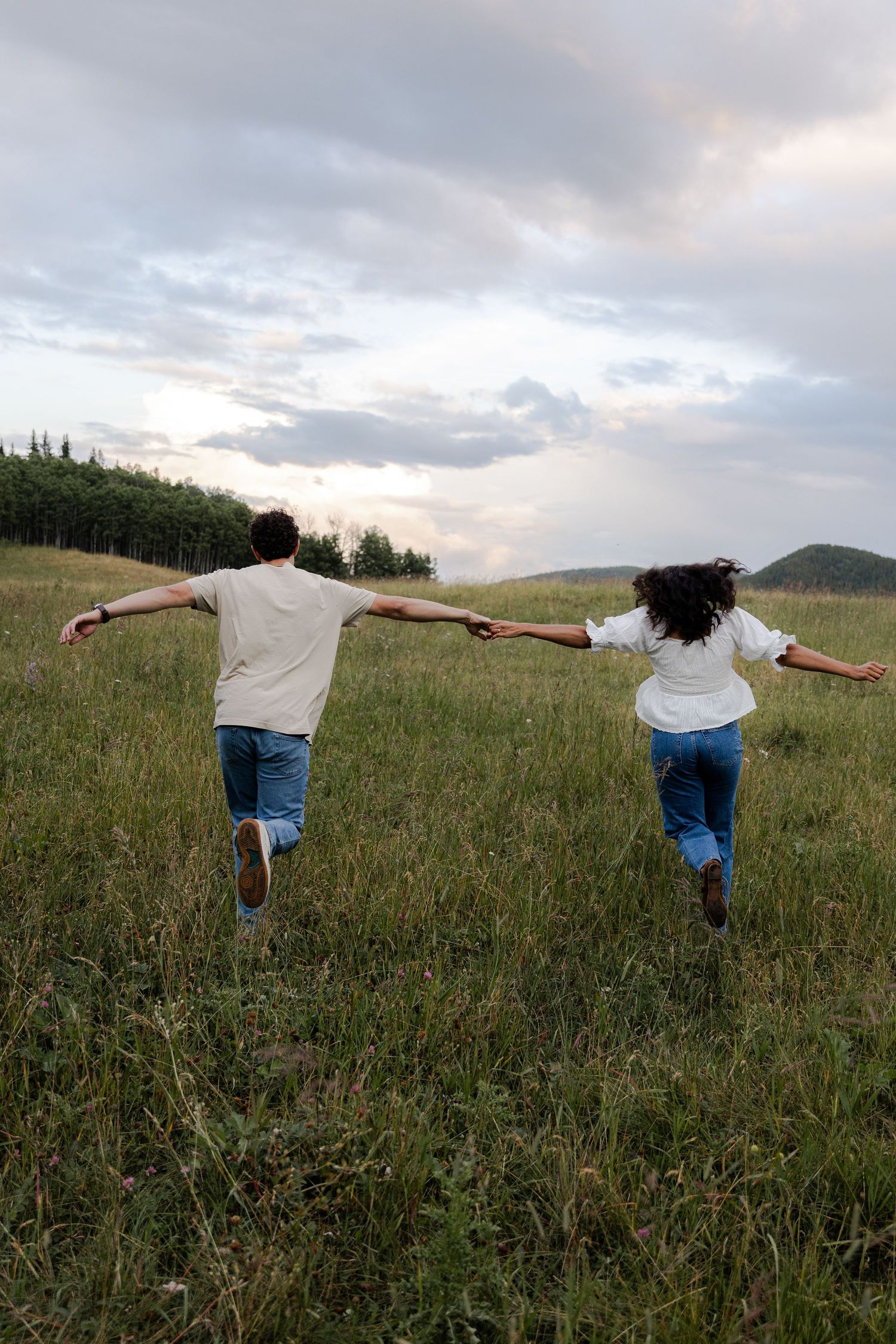 Couple holding hands running through green meadow with hills and cloudy sky in background.
