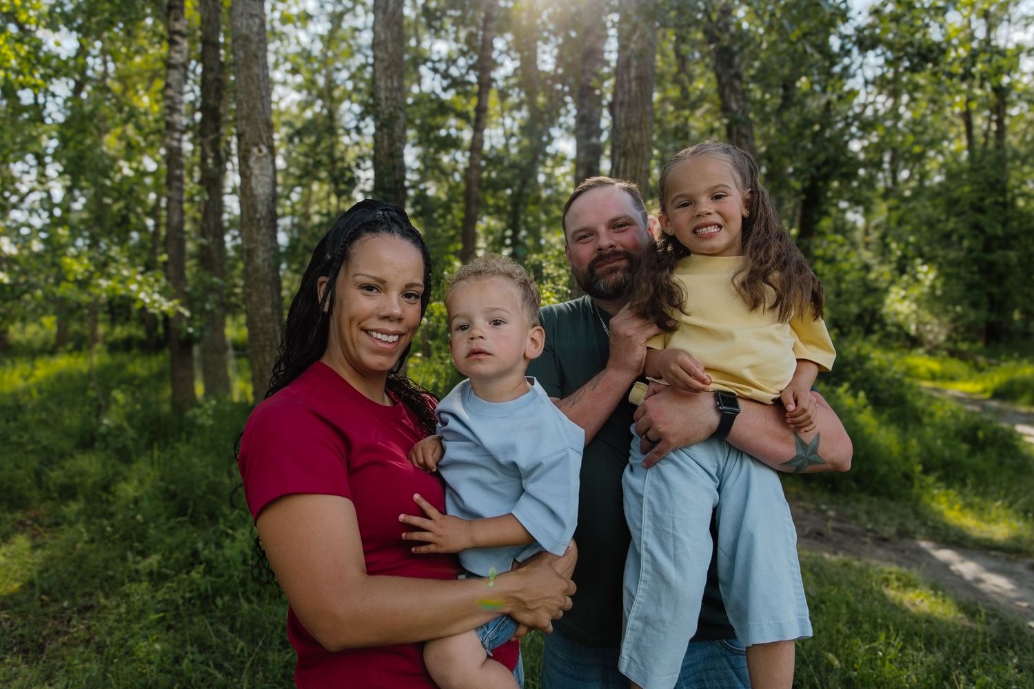 A happy interracial family of four poses outdoors in a lush green forest, parents holding two young children.