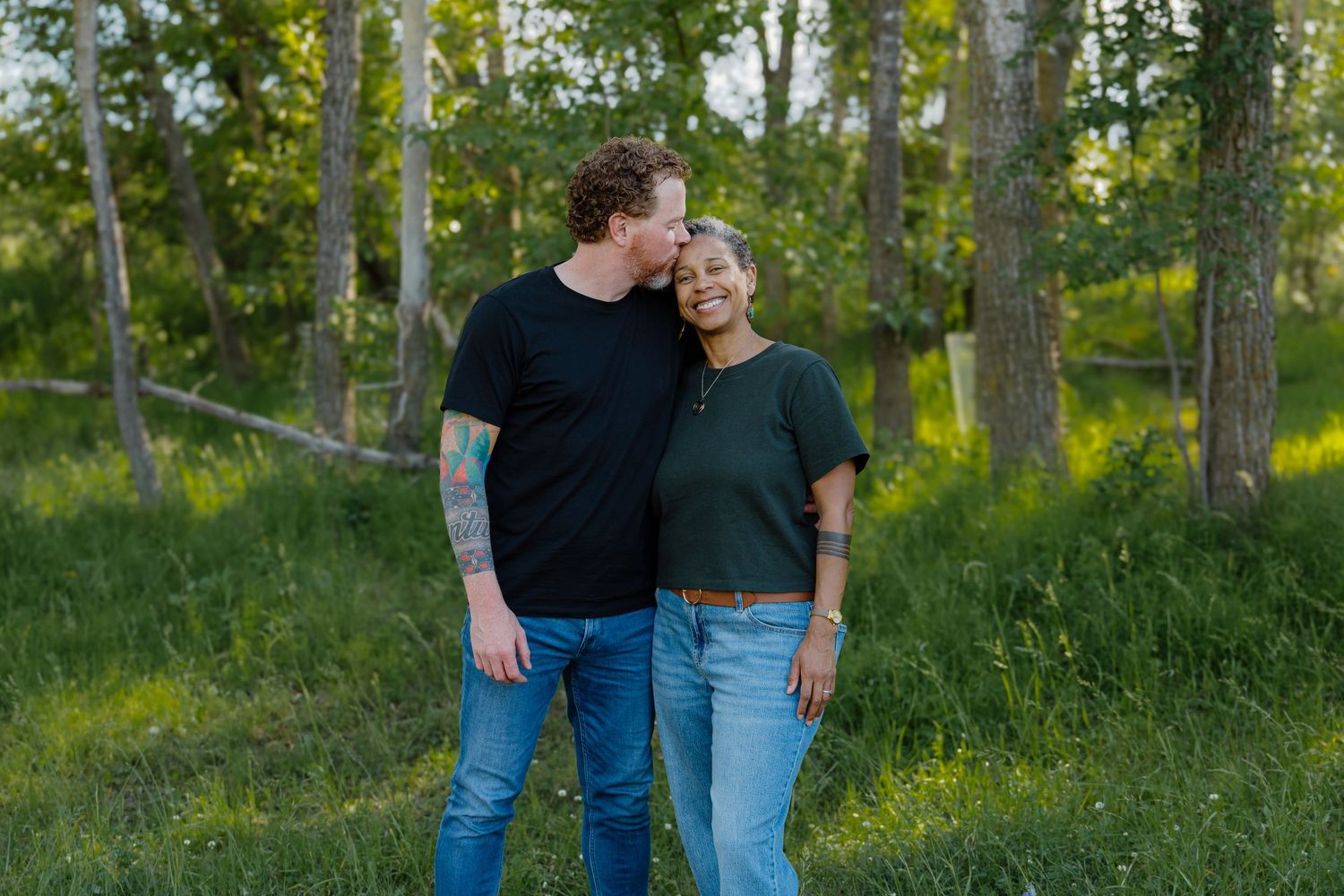 Couple embracing in lush green forest, man kissing woman's temple, both wearing casual jeans and t-shirts outdoors.