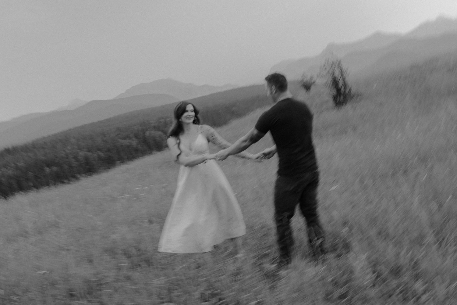 Black and white photo of a couple holding hands and dancing in a grassy mountain meadow.