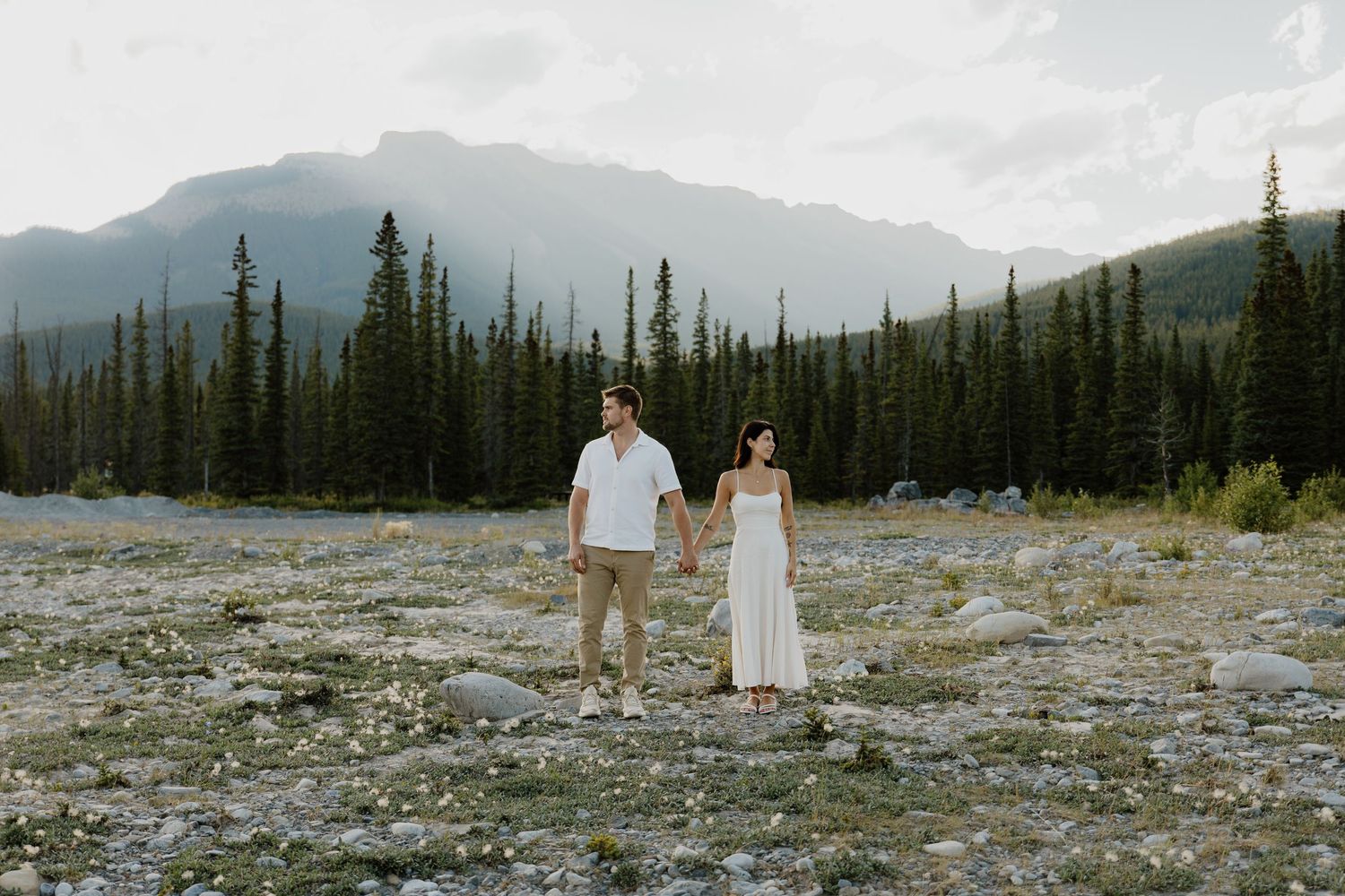 Couple holding hands in white outfits with Rocky Mountain peaks and pine trees in background.