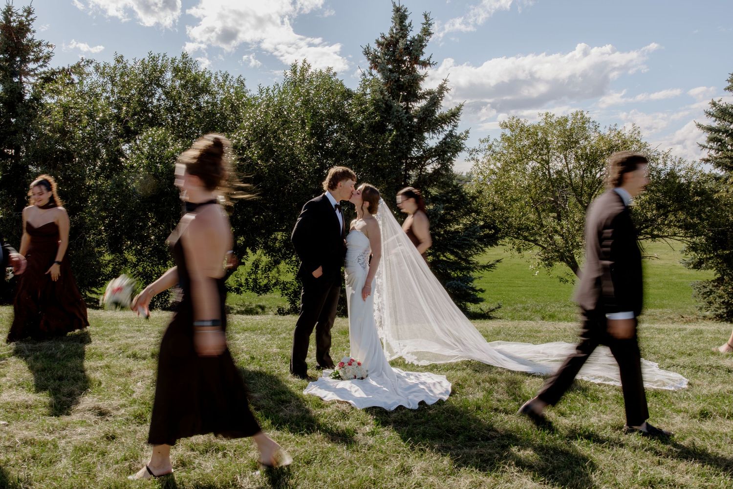 Bride and groom share a kiss while wedding party walks past on a sunny outdoor lawn with green trees.
