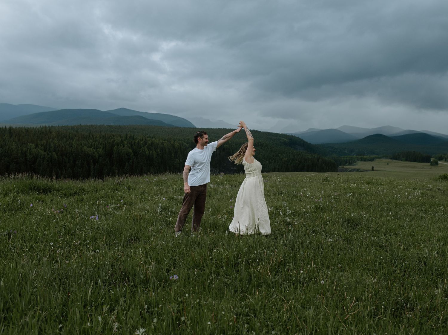 Couple dancing in a green meadow with mountains and cloudy sky backdrop.