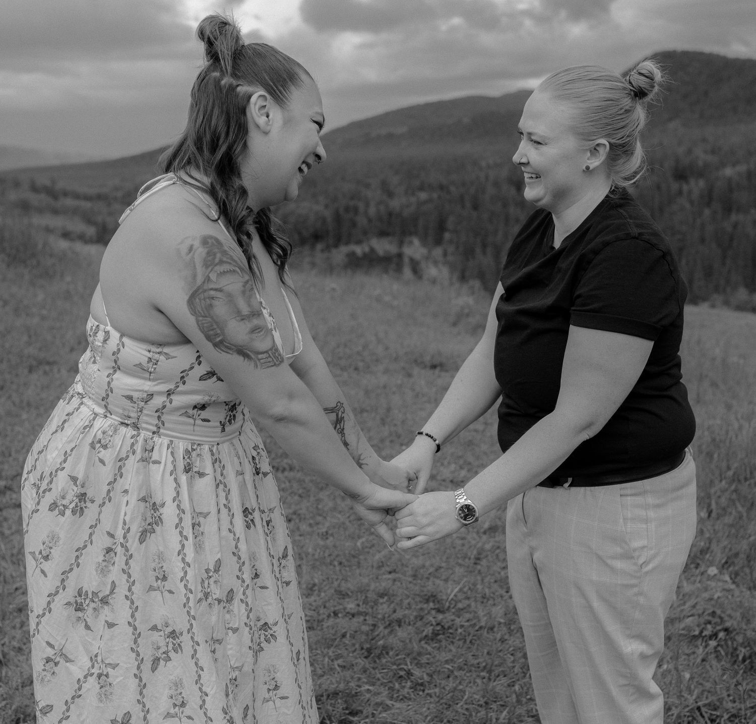 Two women holding hands and smiling at each other in a black and white outdoor portrait.