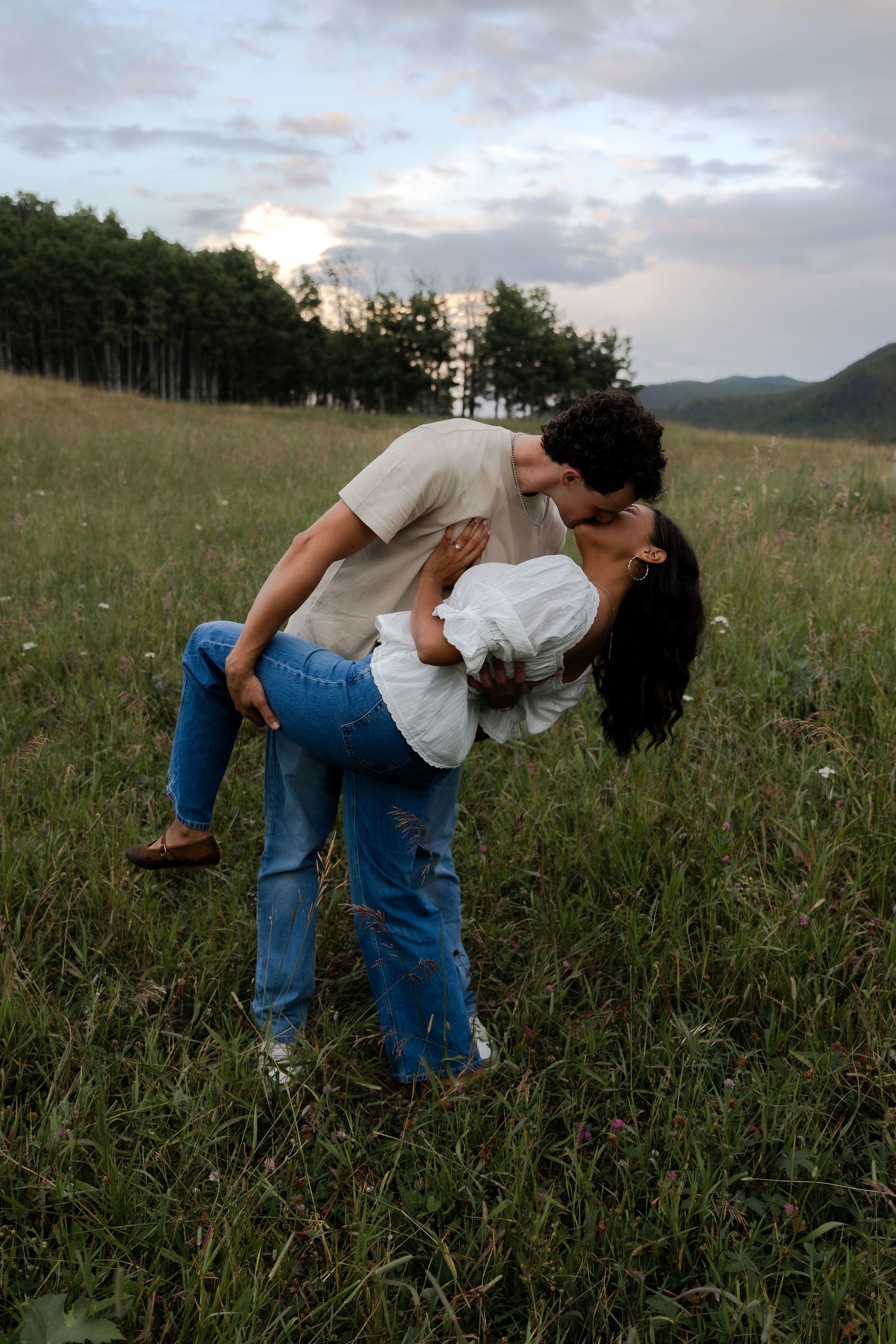 Couple sharing a romantic dip kiss in a green meadow at dusk, with trees and mountains in the background.