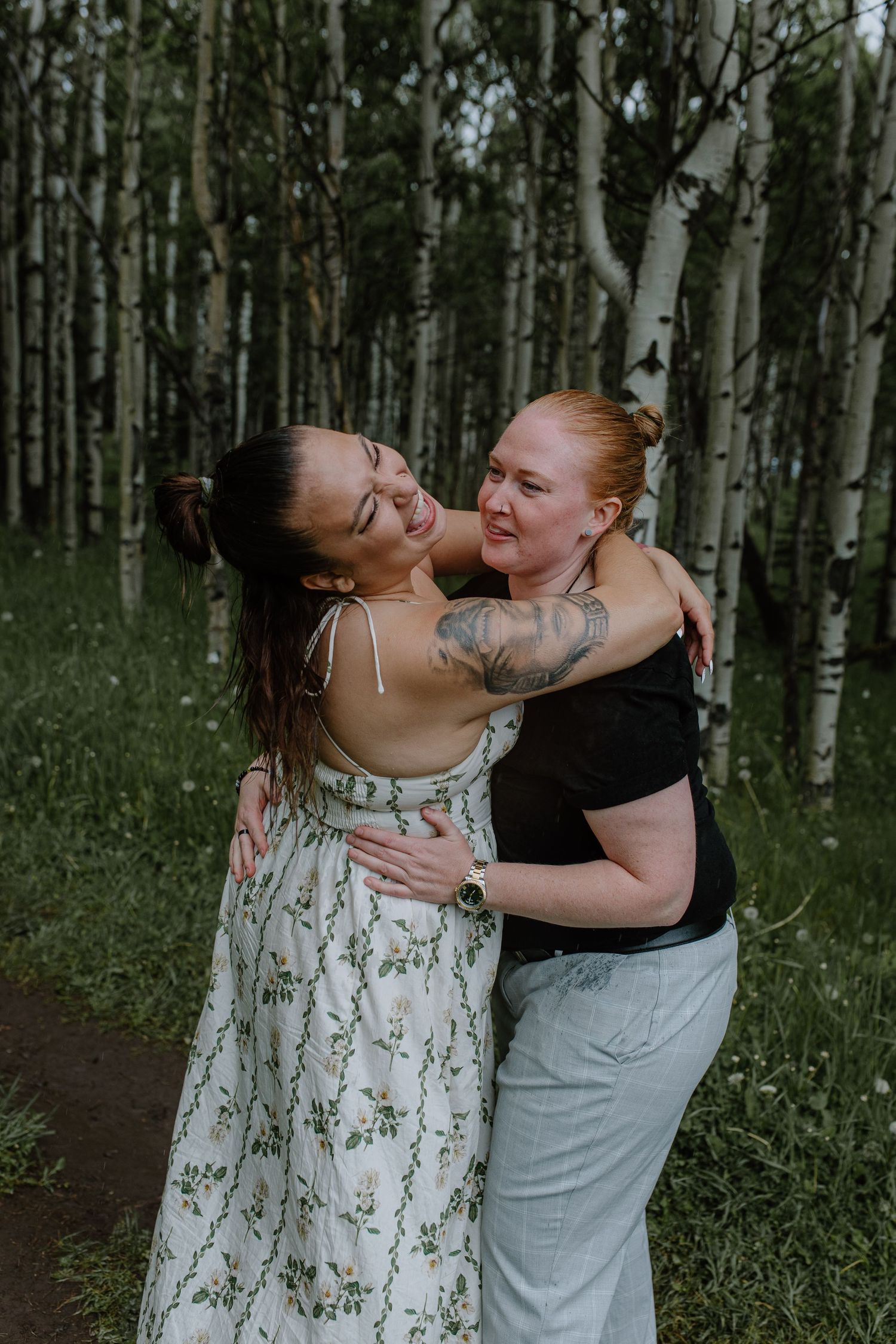 Two women laughing and embracing each other warmly among white birch trees in a forest.