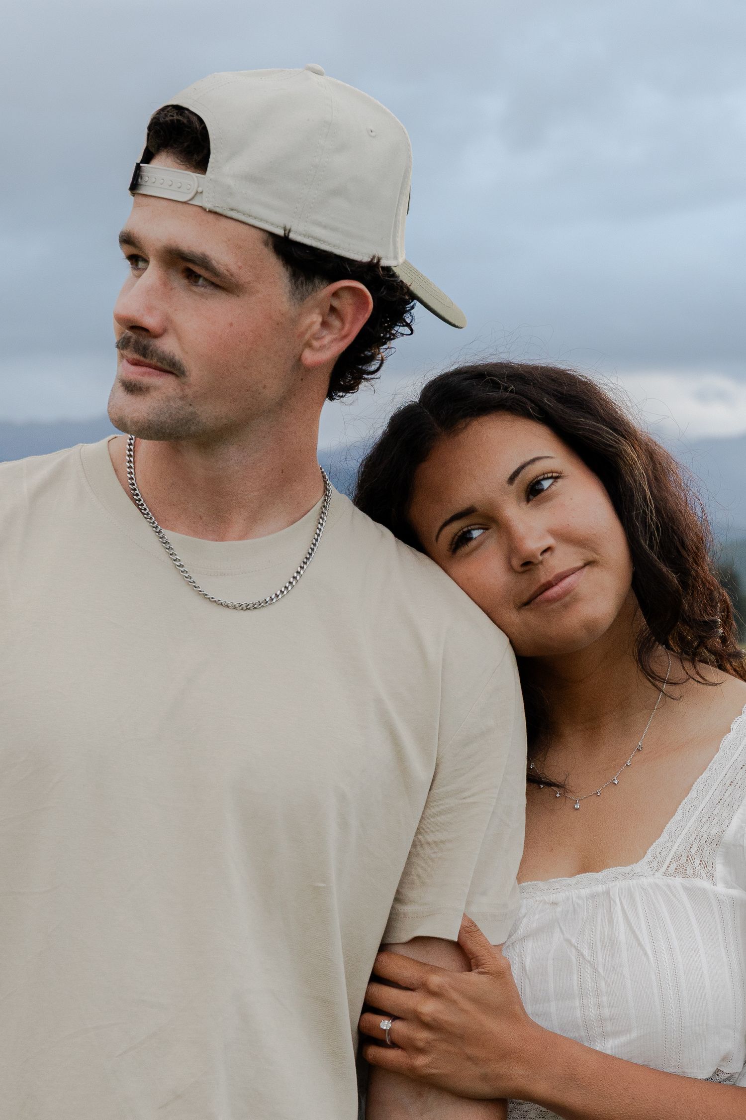 A couple poses outdoors, woman in white dress leaning on man in beige cap and shirt, cloudy sky backdrop.