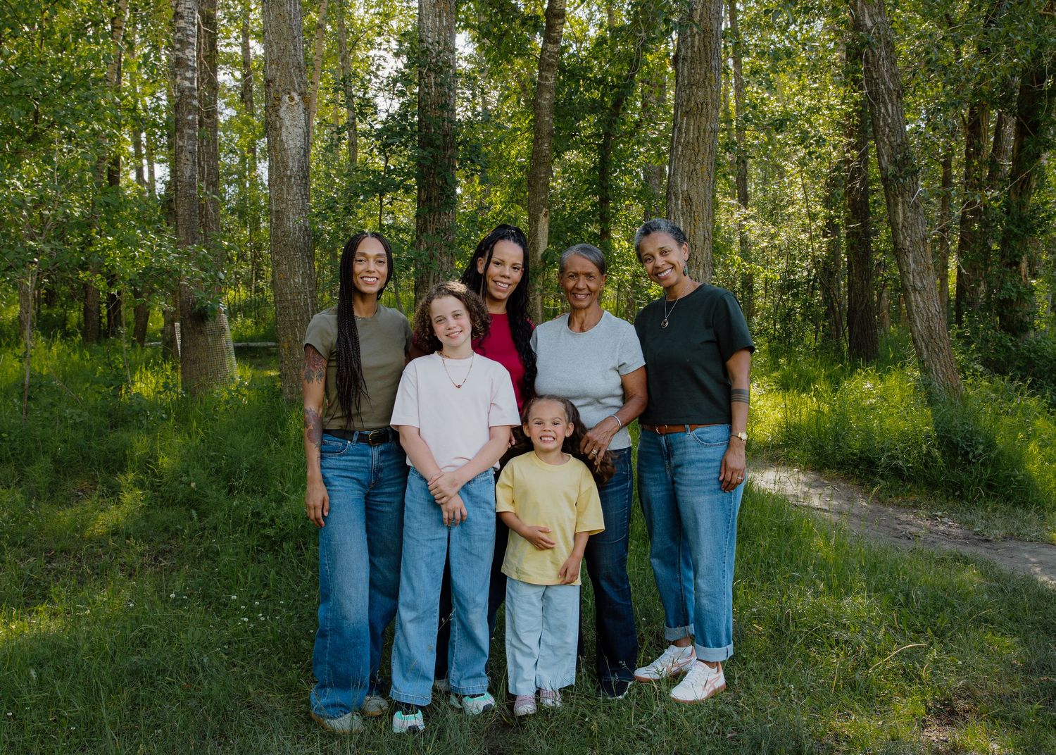 Multi-generational family group smiling together on a lush green forest trail surrounded by tall trees.