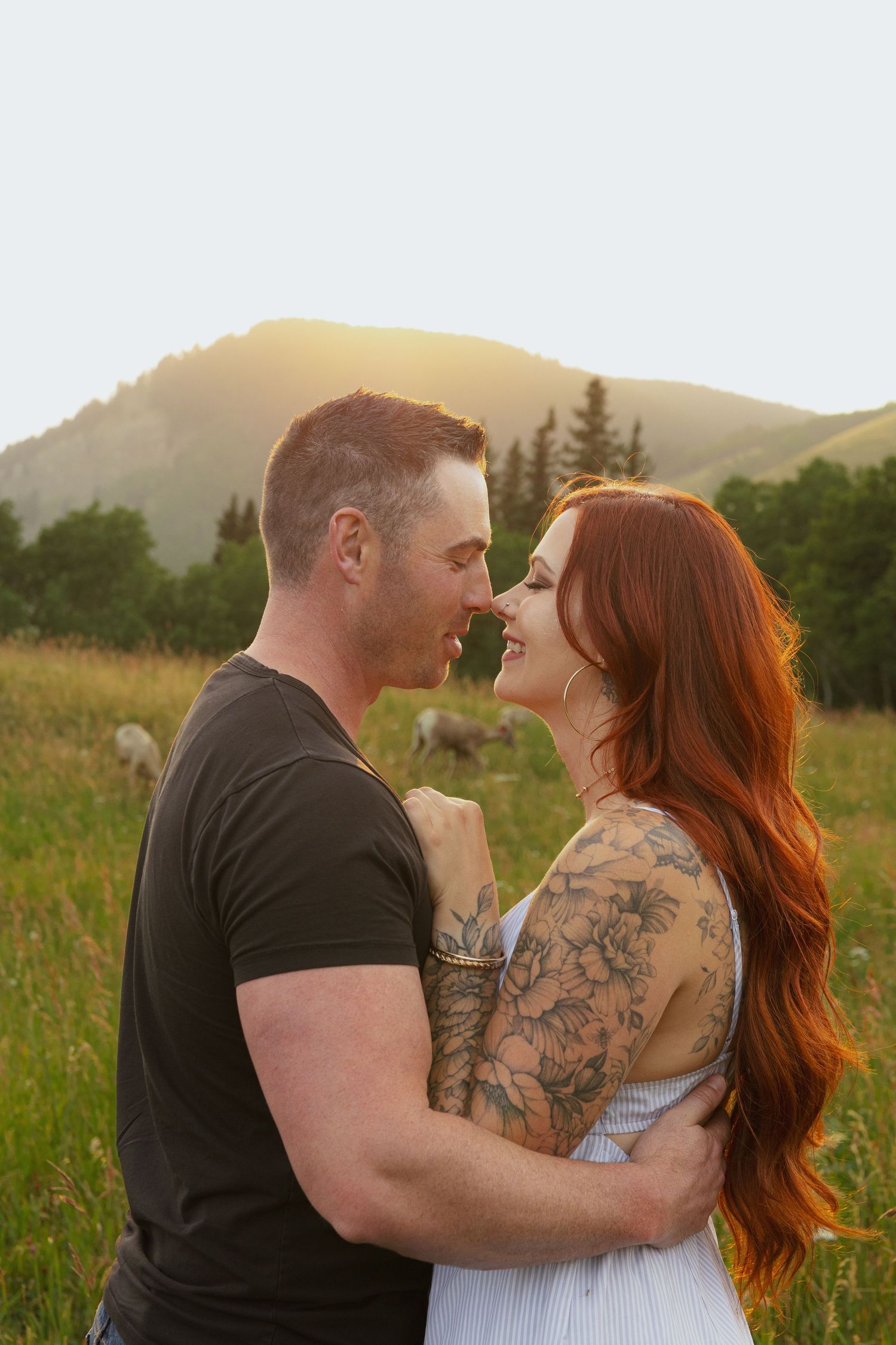 A couple with tattoos share a romantic moment in a sunlit mountain meadow at golden hour.