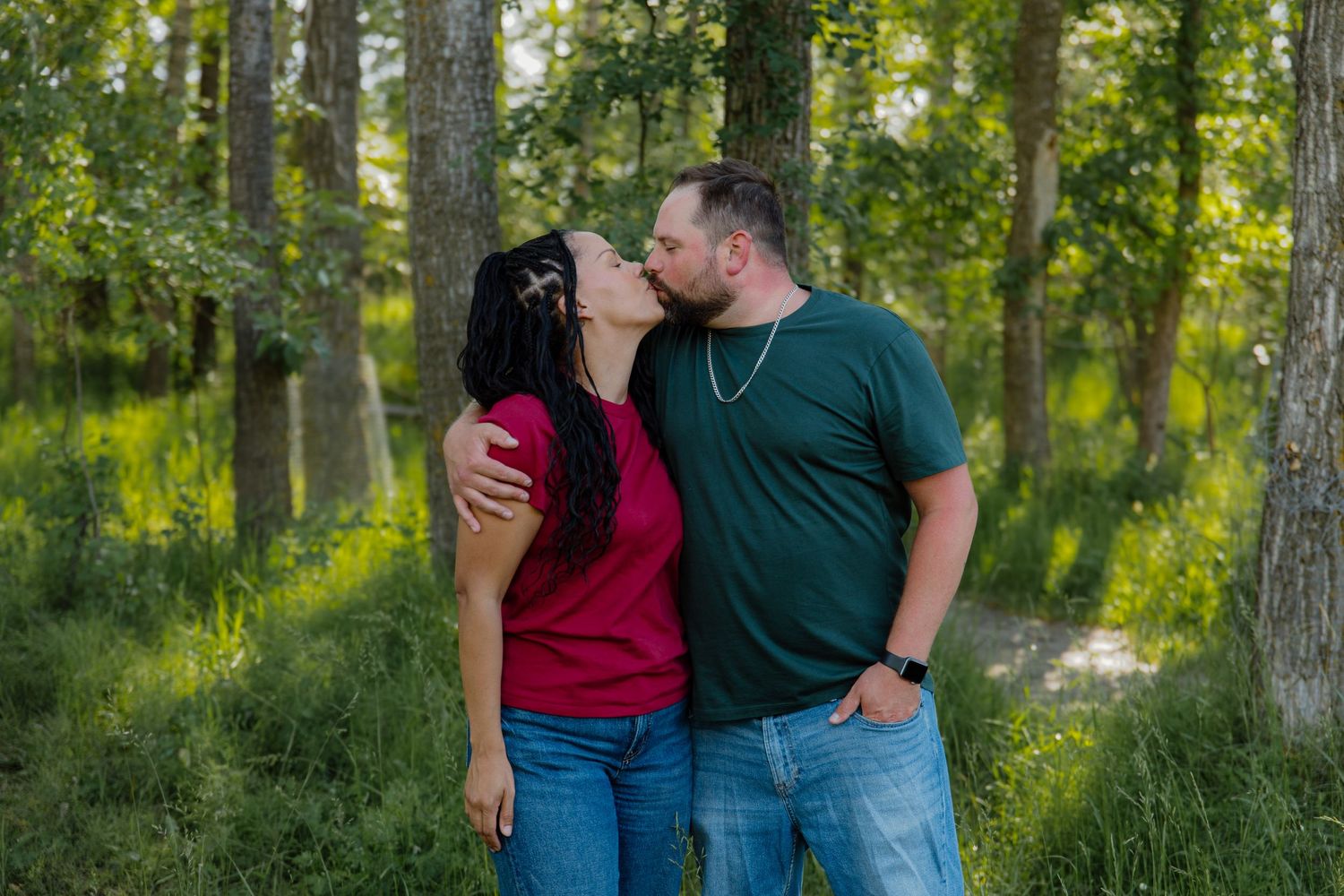 A couple shares a romantic kiss in a lush green forest, wearing casual red and green shirts with jeans.
