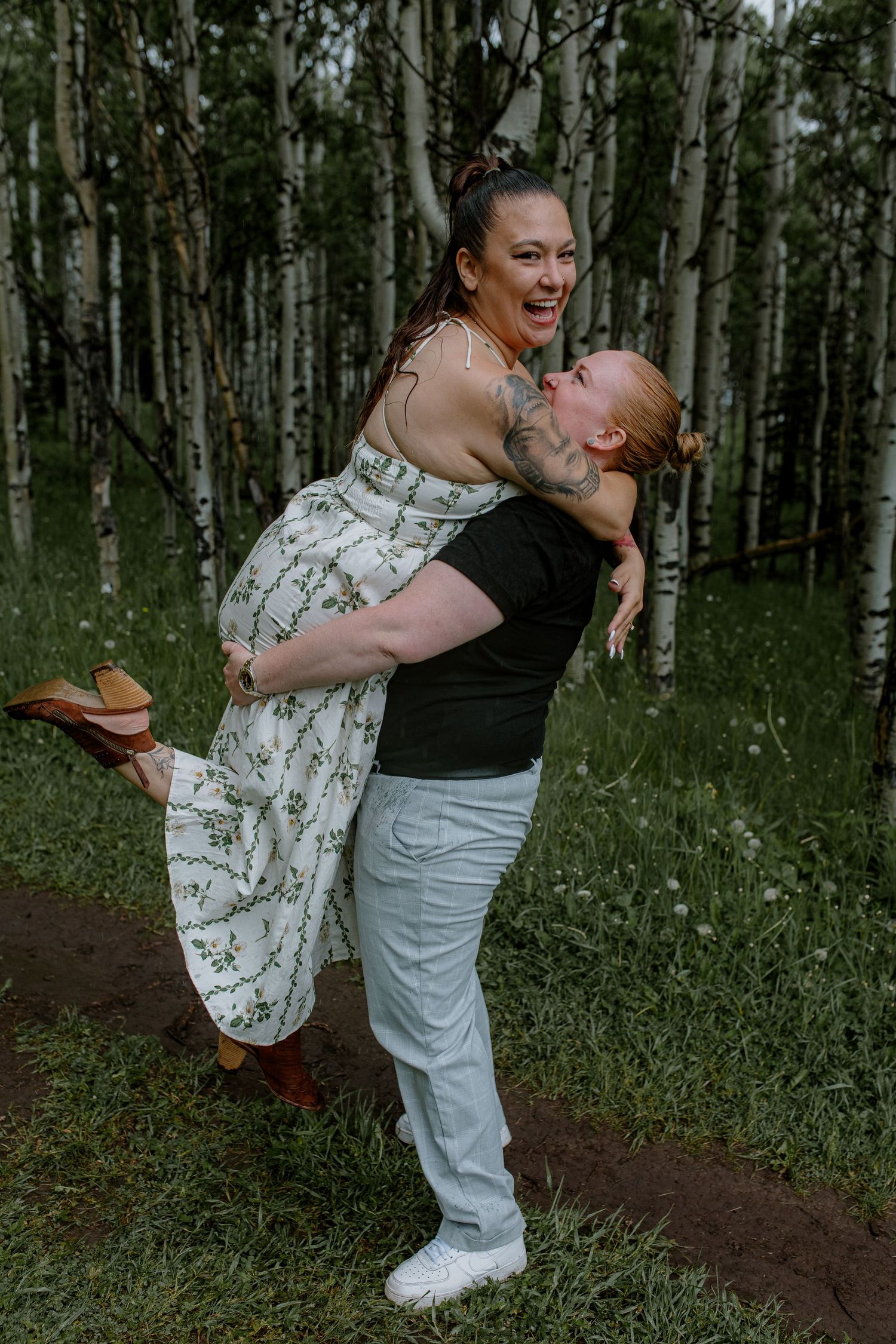 A woman laughs joyfully as her partner lifts her up in a sunlit aspen forest during an engagement photo session.
