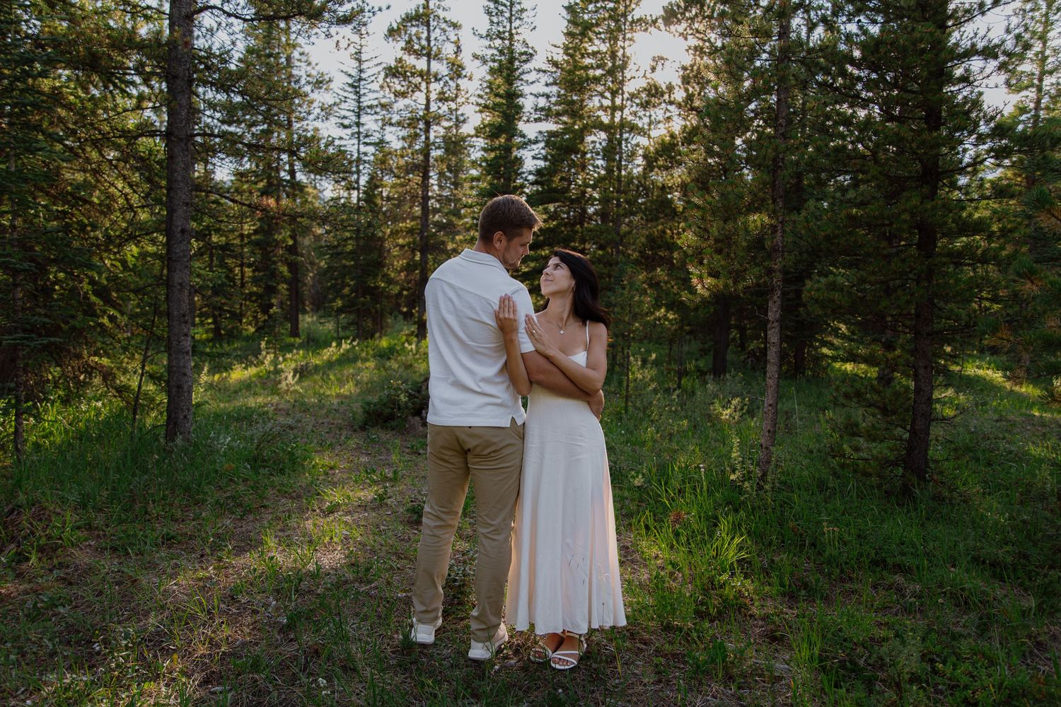 Couple embracing in a lush forest clearing at golden hour, woman in white dress, man in casual attire.