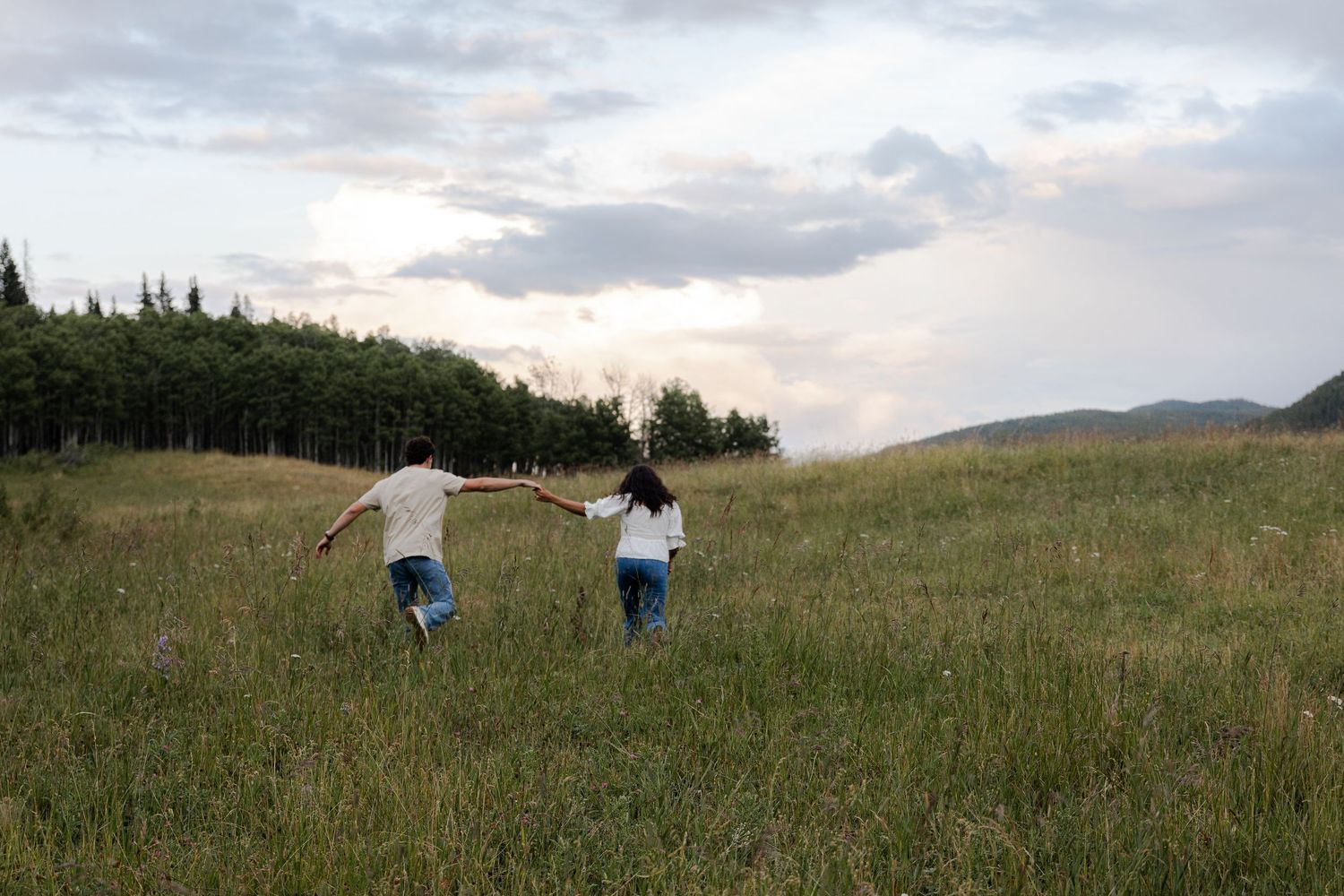 Couple holding hands running through a lush green meadow with trees and cloudy sky in background.