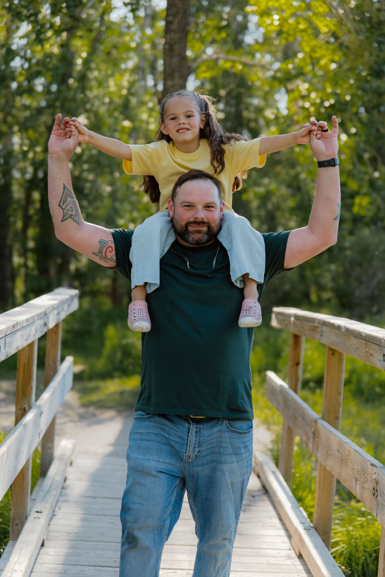 Father carrying young daughter on shoulders on wooden bridge surrounded by green trees.