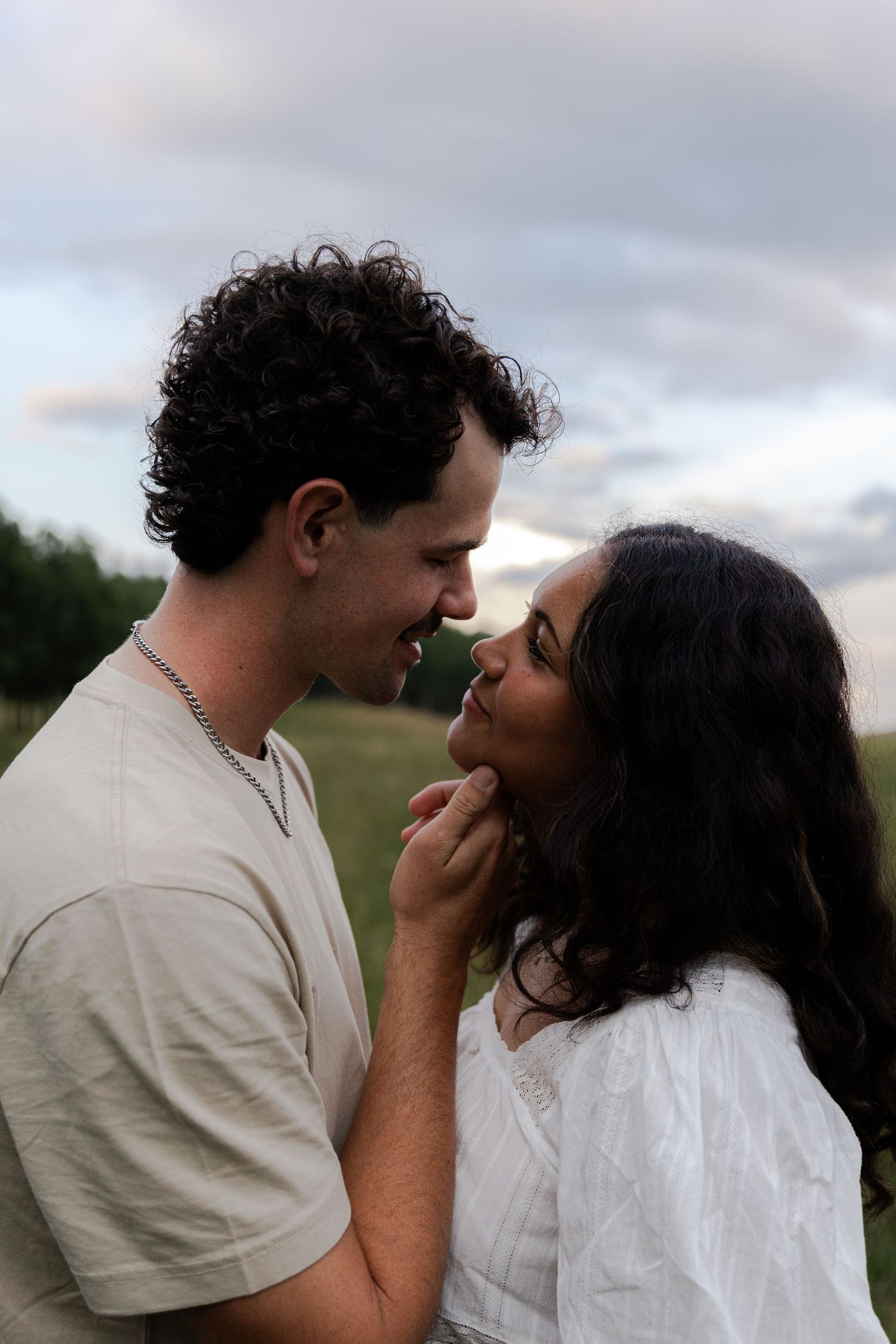 A romantic couple sharing a tender moment outdoors, the man gently touching the woman's chin under a cloudy sky.