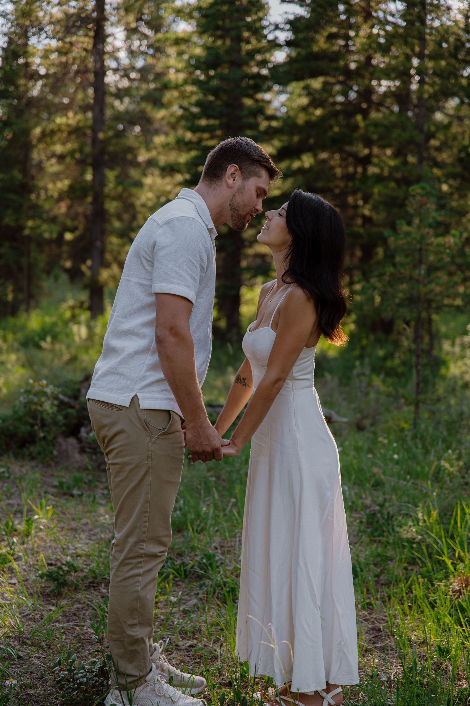 Couple holding hands and sharing a romantic moment in a lush forest clearing, woman in white dress.