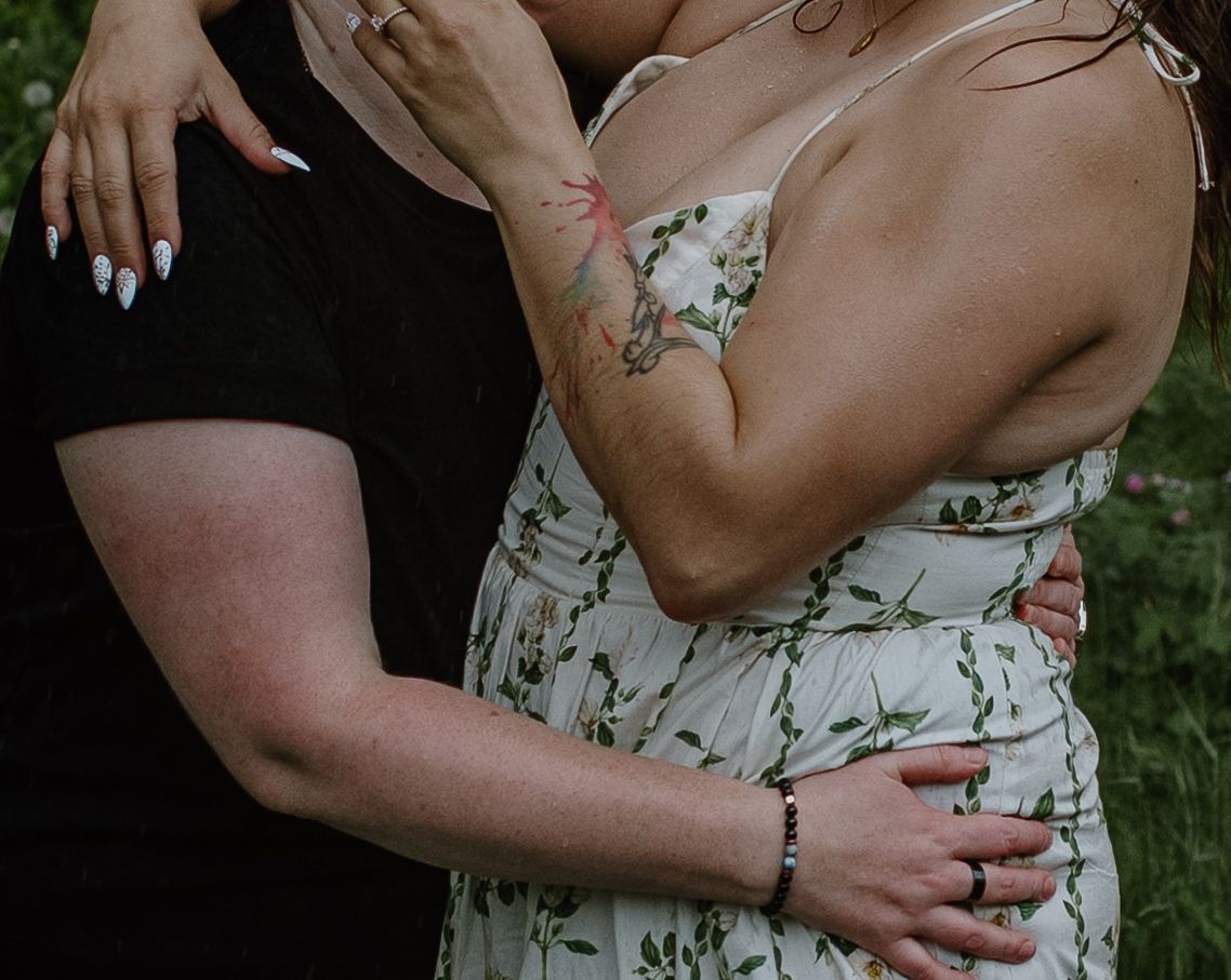 Two women in an intimate embrace, one in a floral dress with a tattoo, the other in a black shirt, outdoors.