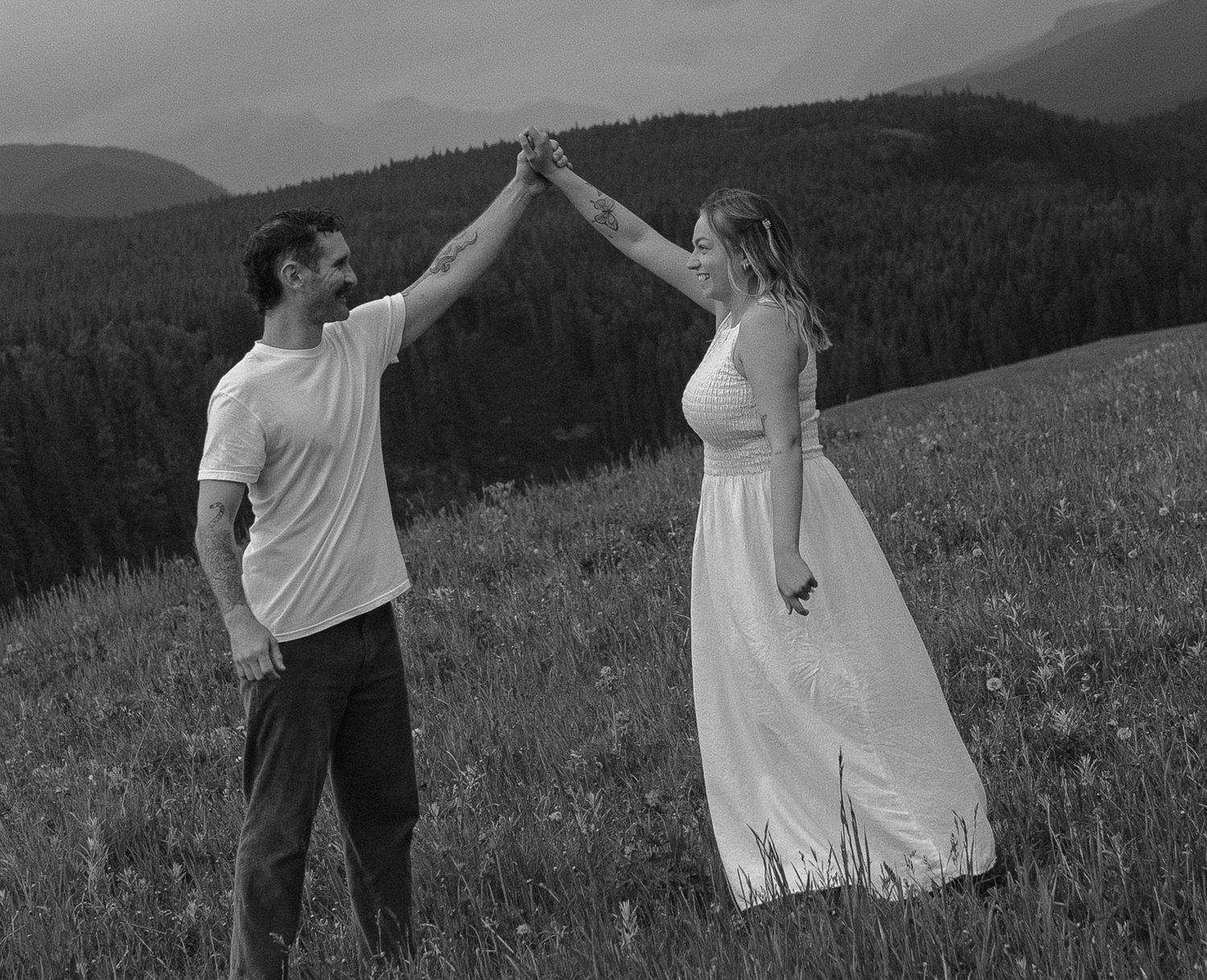 Black and white photo of a couple dancing in a mountain meadow, man in white shirt, woman in flowing white dress.