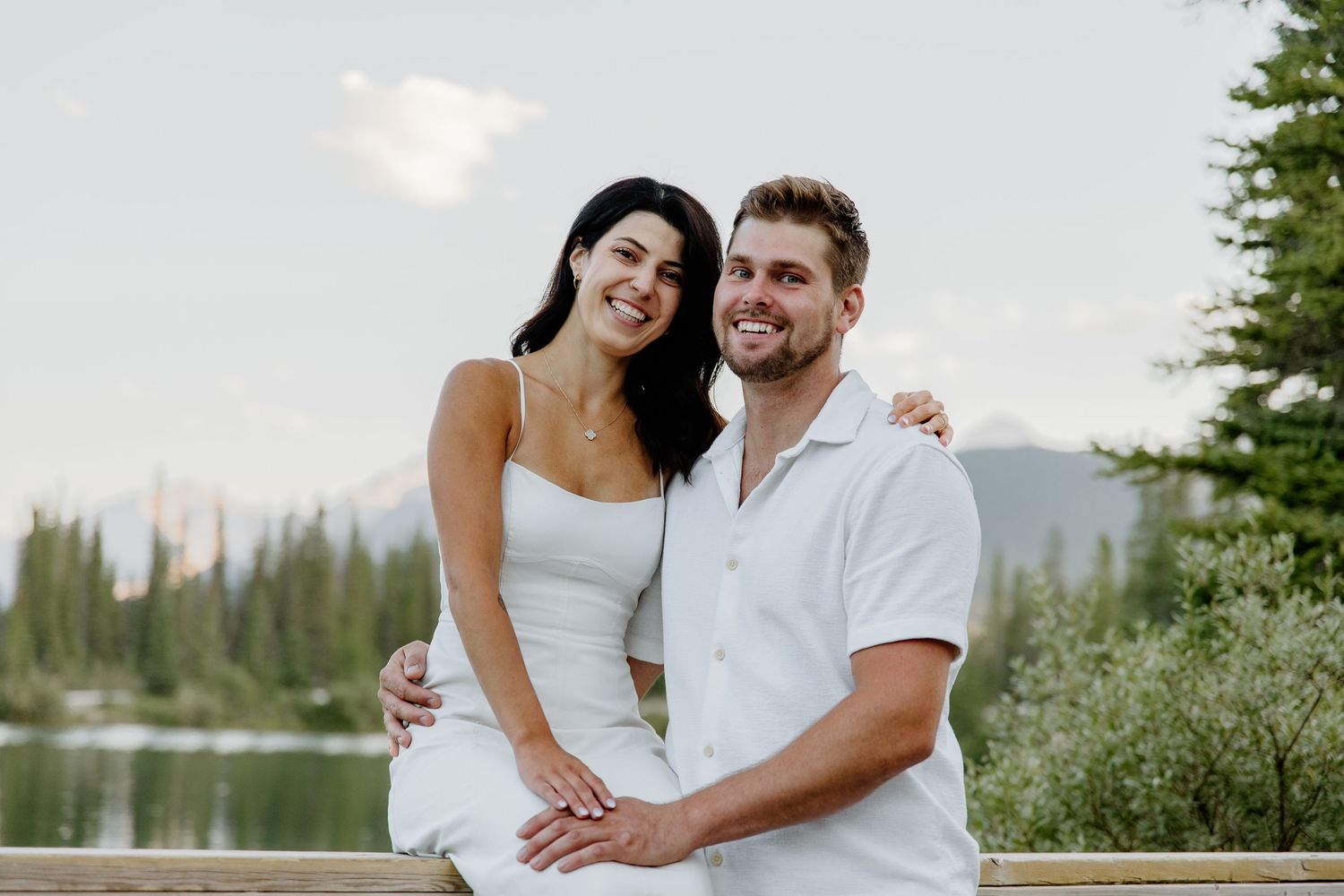 Smiling couple in white outfits posing by a mountain lake surrounded by pine trees.