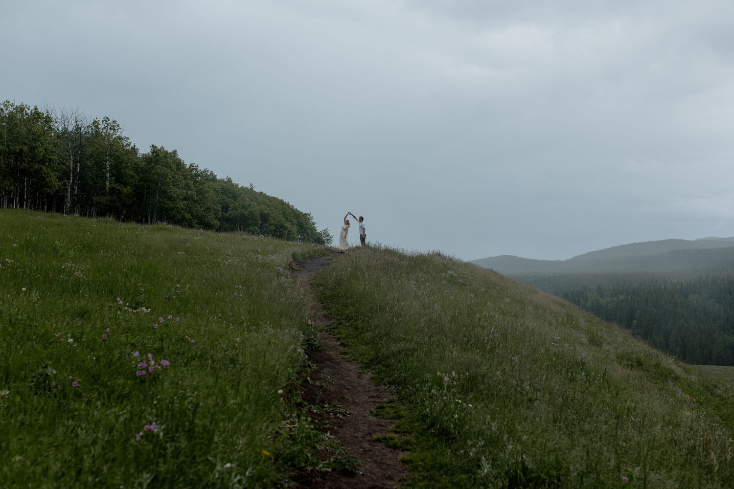 Two people standing on a grassy hilltop trail with mountain forest views under overcast skies.