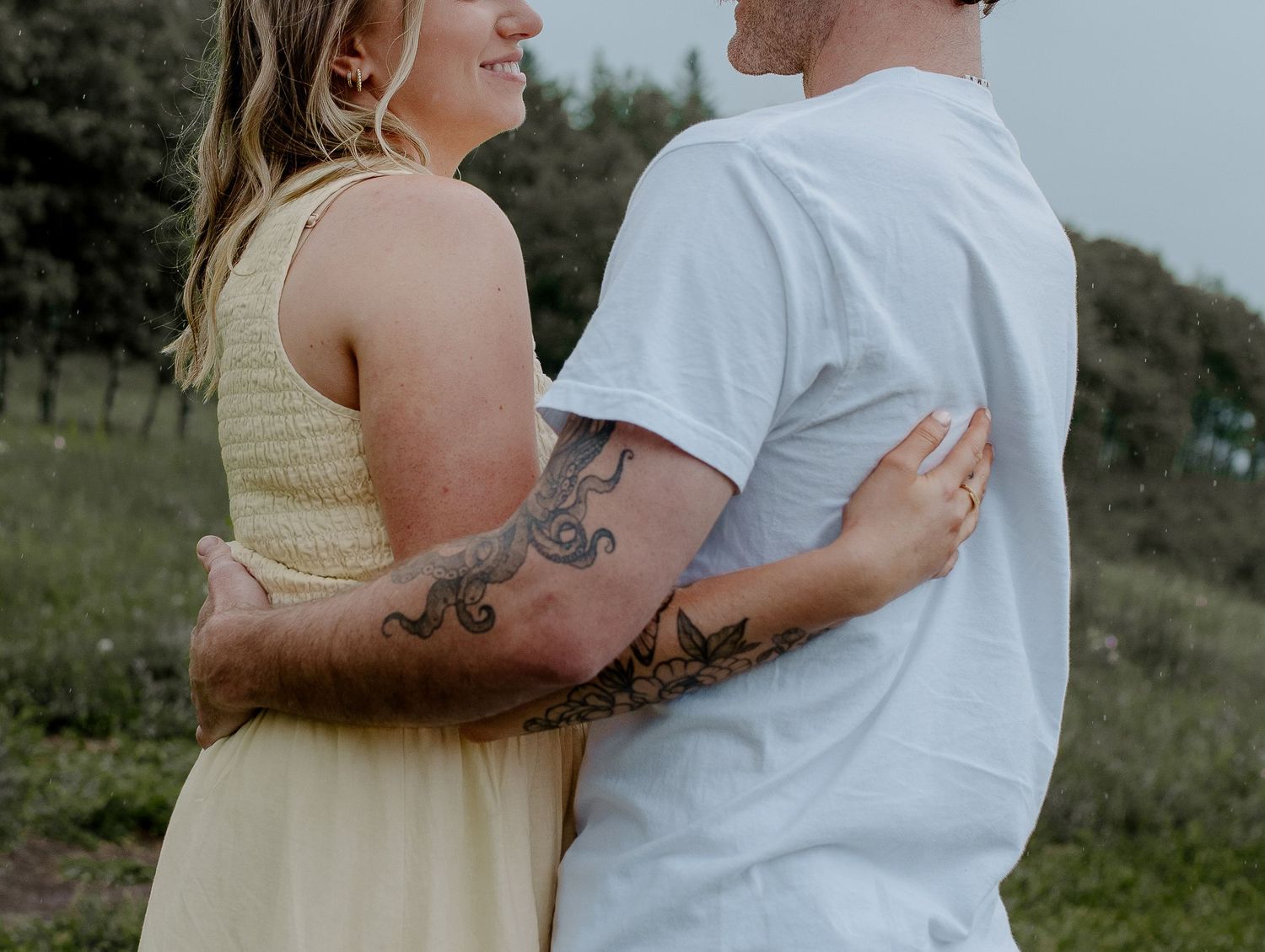 Couple embracing outdoors, man with tattoos wearing white t-shirt and woman in yellow dress.