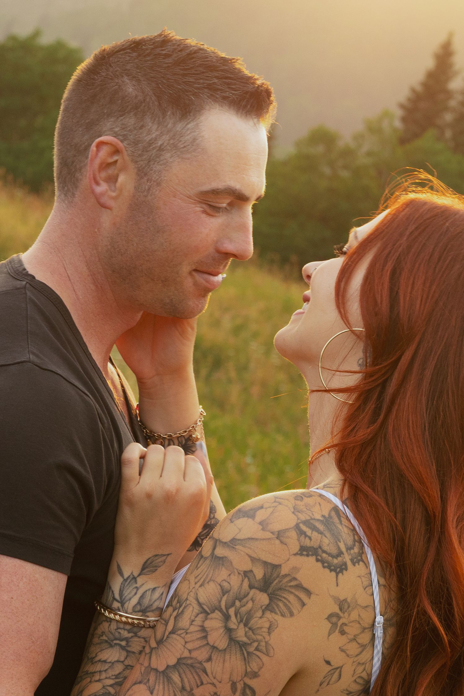Couple sharing a romantic moment outdoors at golden hour, woman with red hair and floral tattoos, man in black t-shirt.