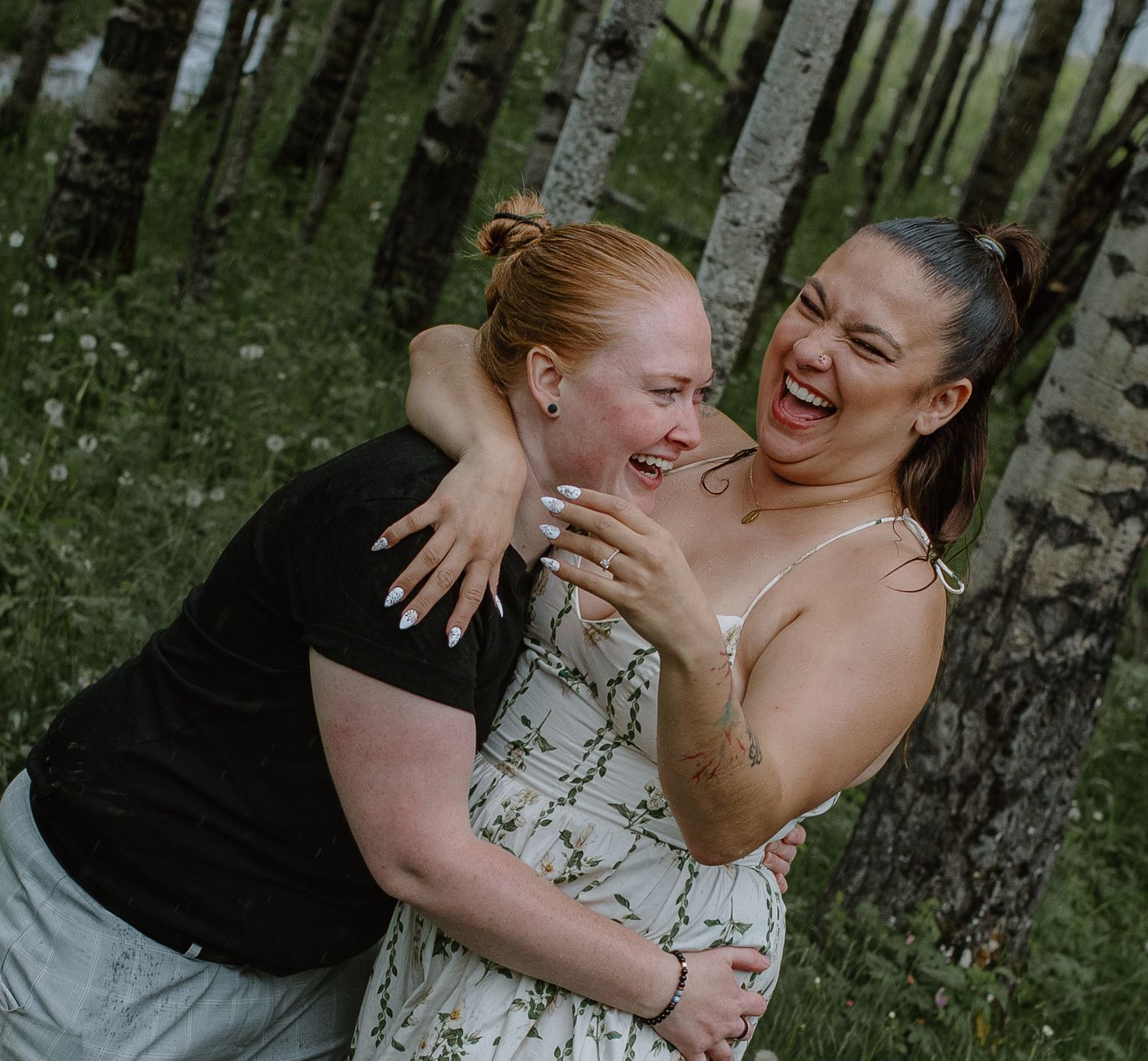 Two women laughing and hugging joyfully among white birch trees in a lush green forest.
