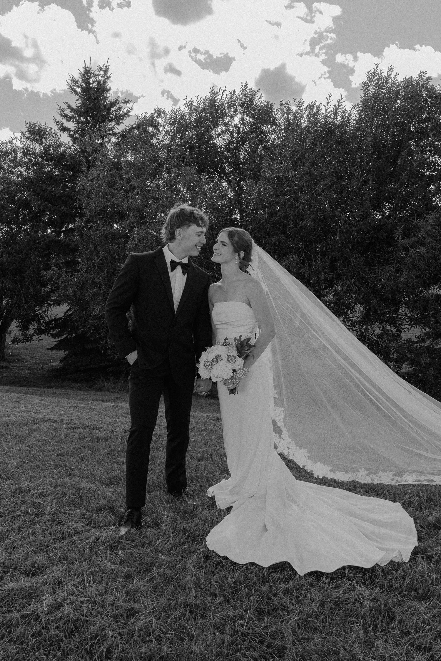 Black and white photo of a bride and groom gazing at each other outdoors, her veil flowing in the wind.
