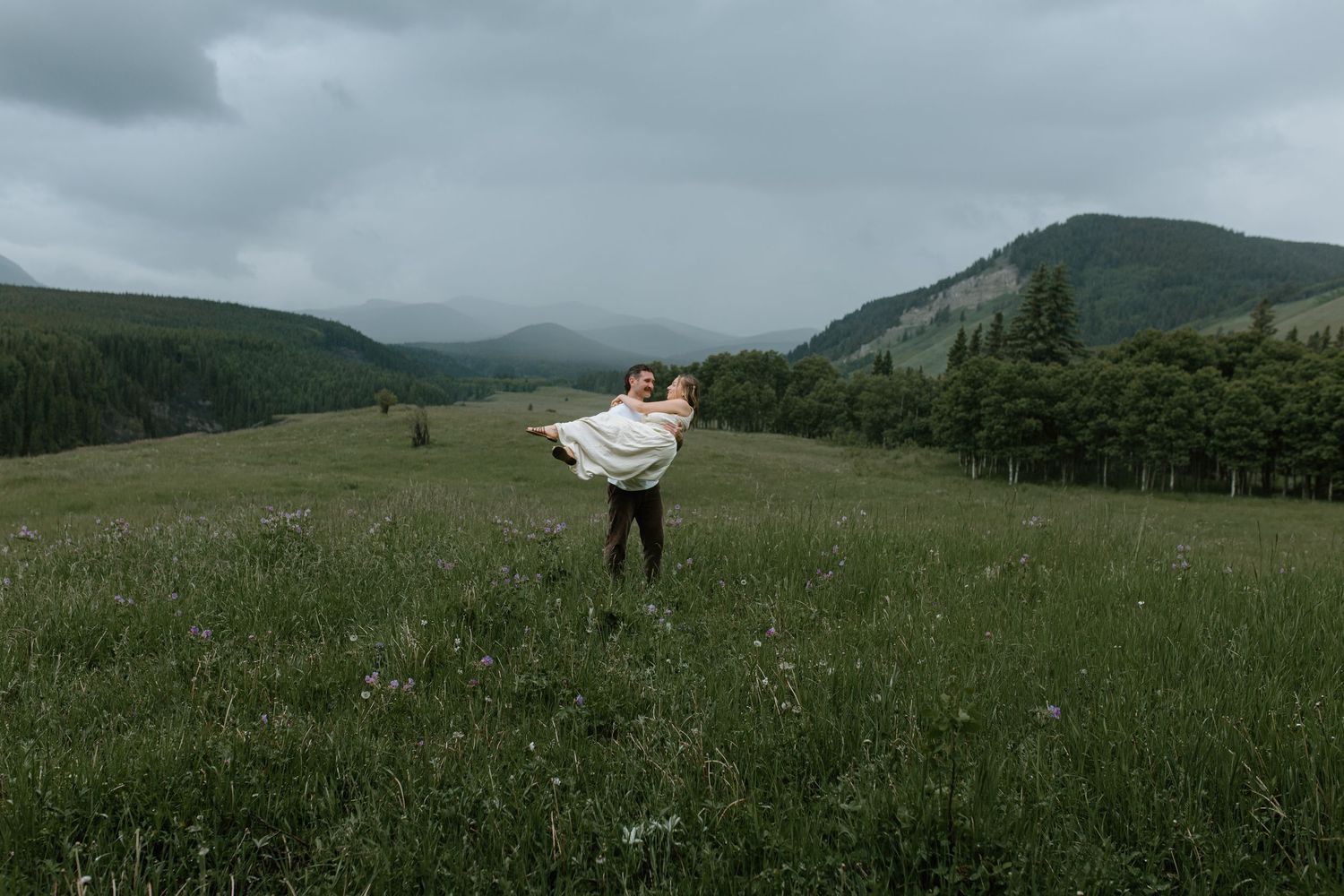 Man lifting woman in a green meadow with forested mountains and dramatic cloudy sky in the background.