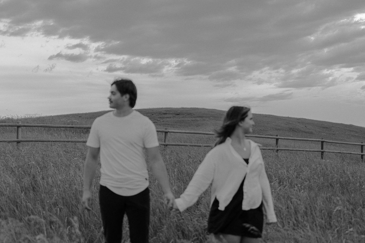 Black and white photo of a couple holding hands in a field with a fence and cloudy sky backdrop.