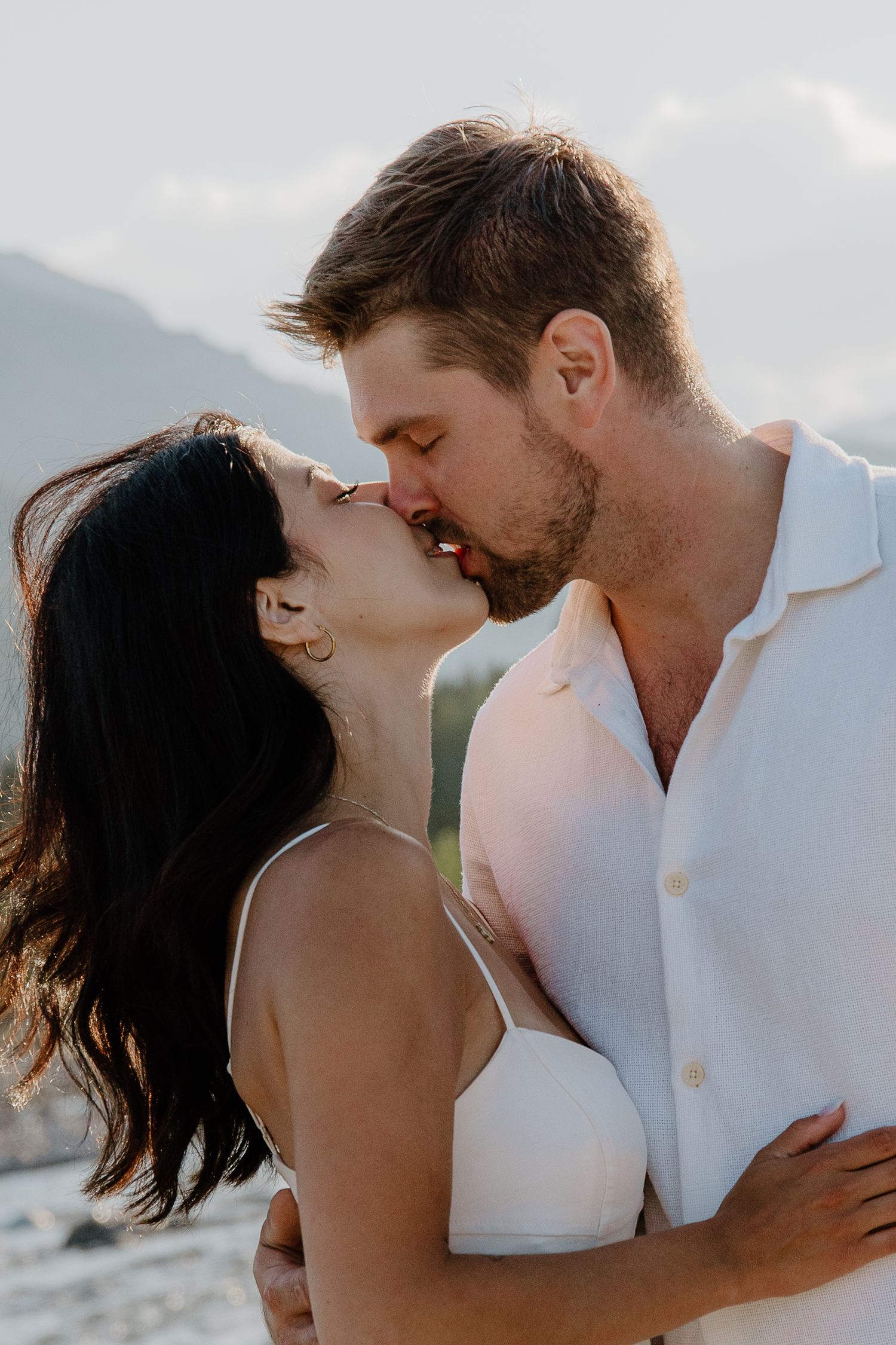 A couple shares a romantic kiss outdoors with mountains in the background, both dressed in white.