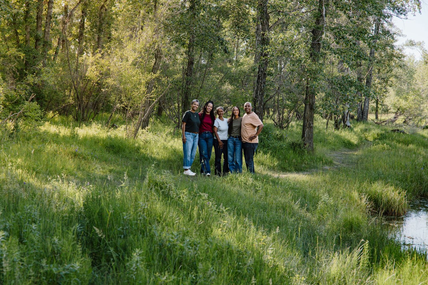 Family of five posing together in a lush green meadow surrounded by tall trees and a small stream.