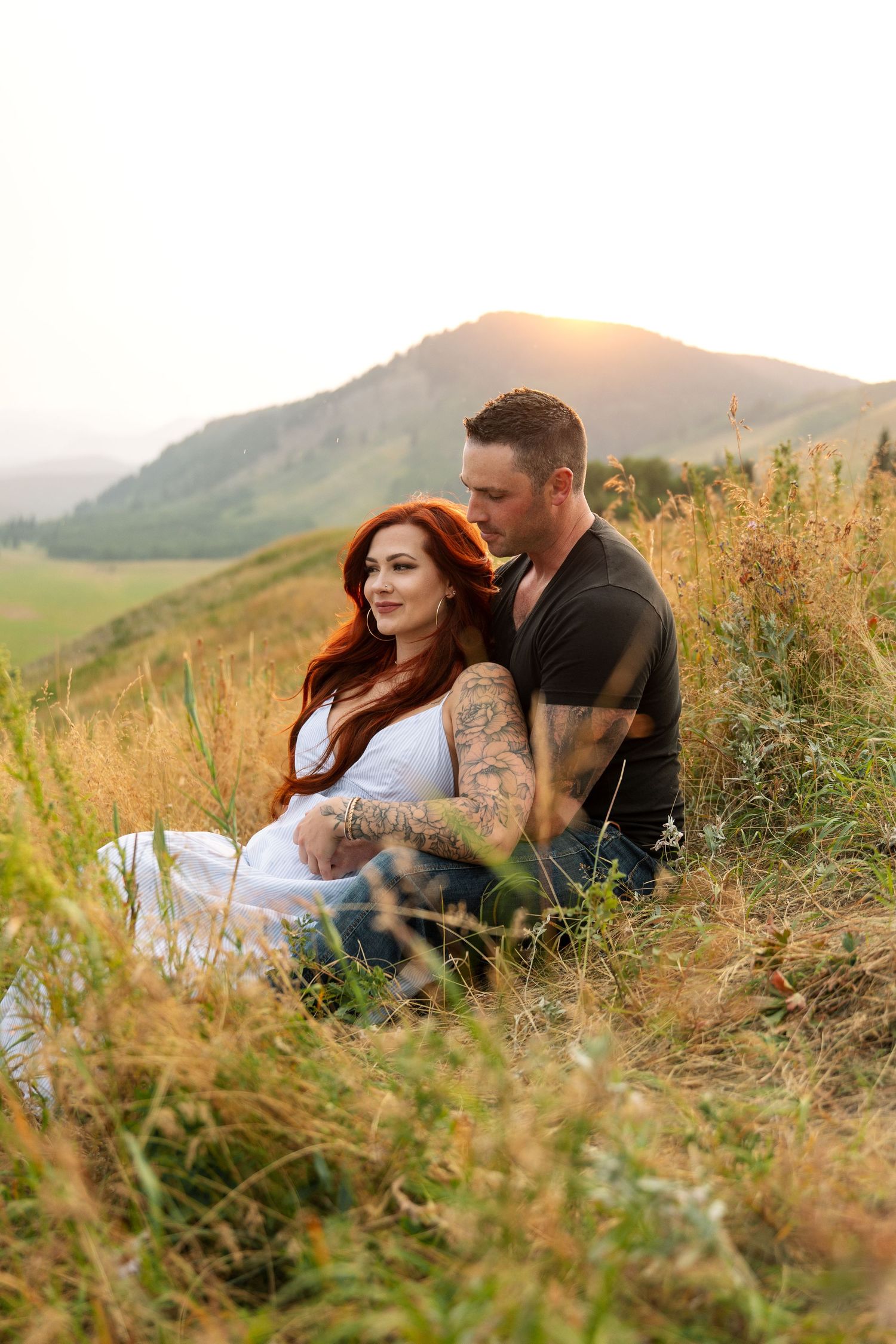 Couple sitting together in golden hour sunlight on a grassy hillside, woman in white dress with red hair, man in black shirt.