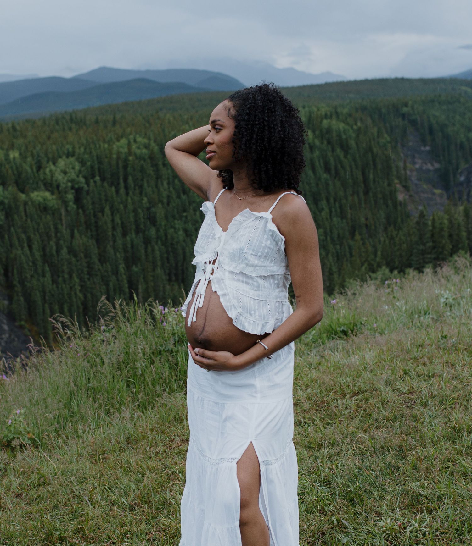 Pregnant woman in white dress posing on a mountain hillside surrounded by lush green forest.