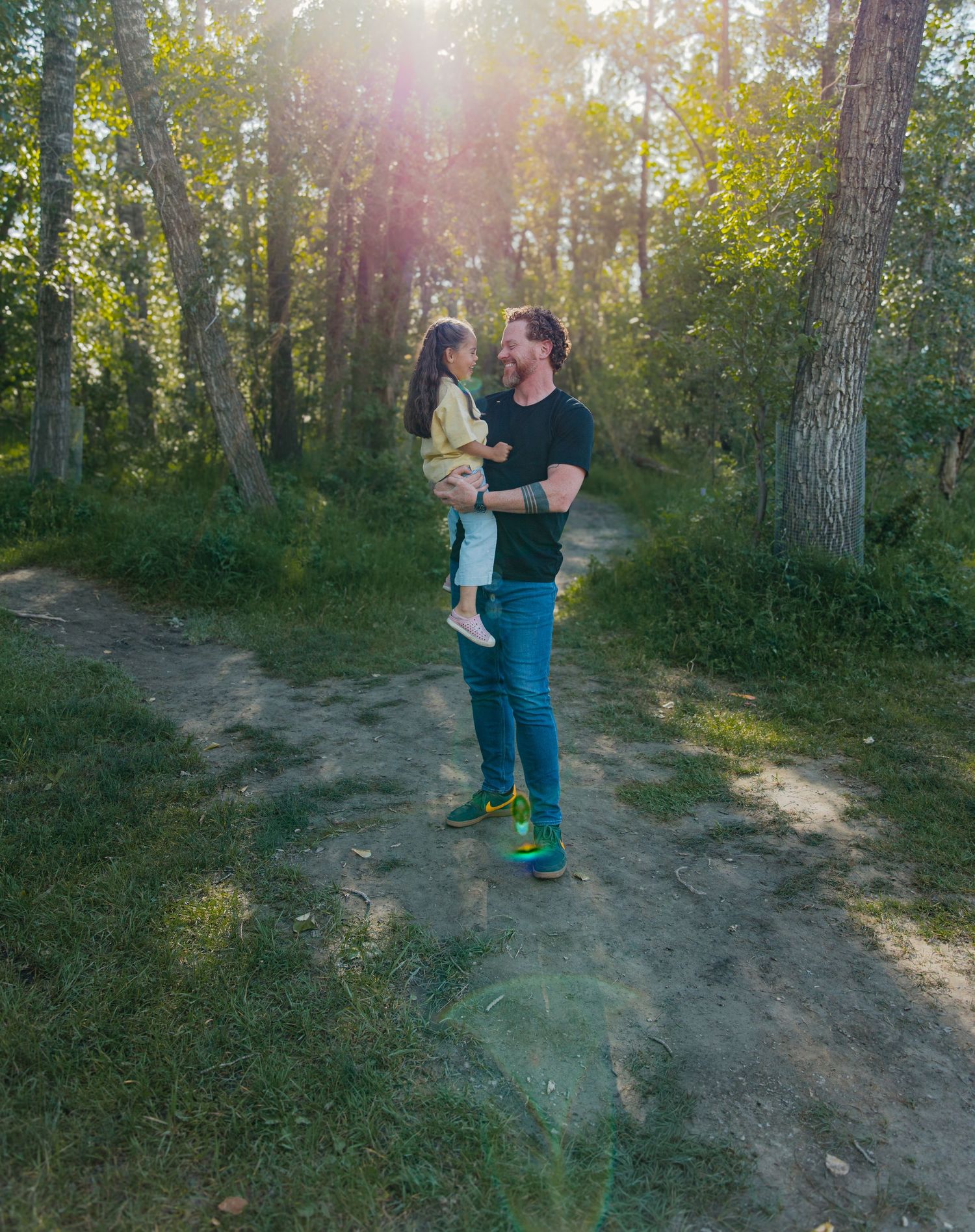A man holds a young child in a sunlit forest clearing, both smiling warmly among green trees on a dirt path.